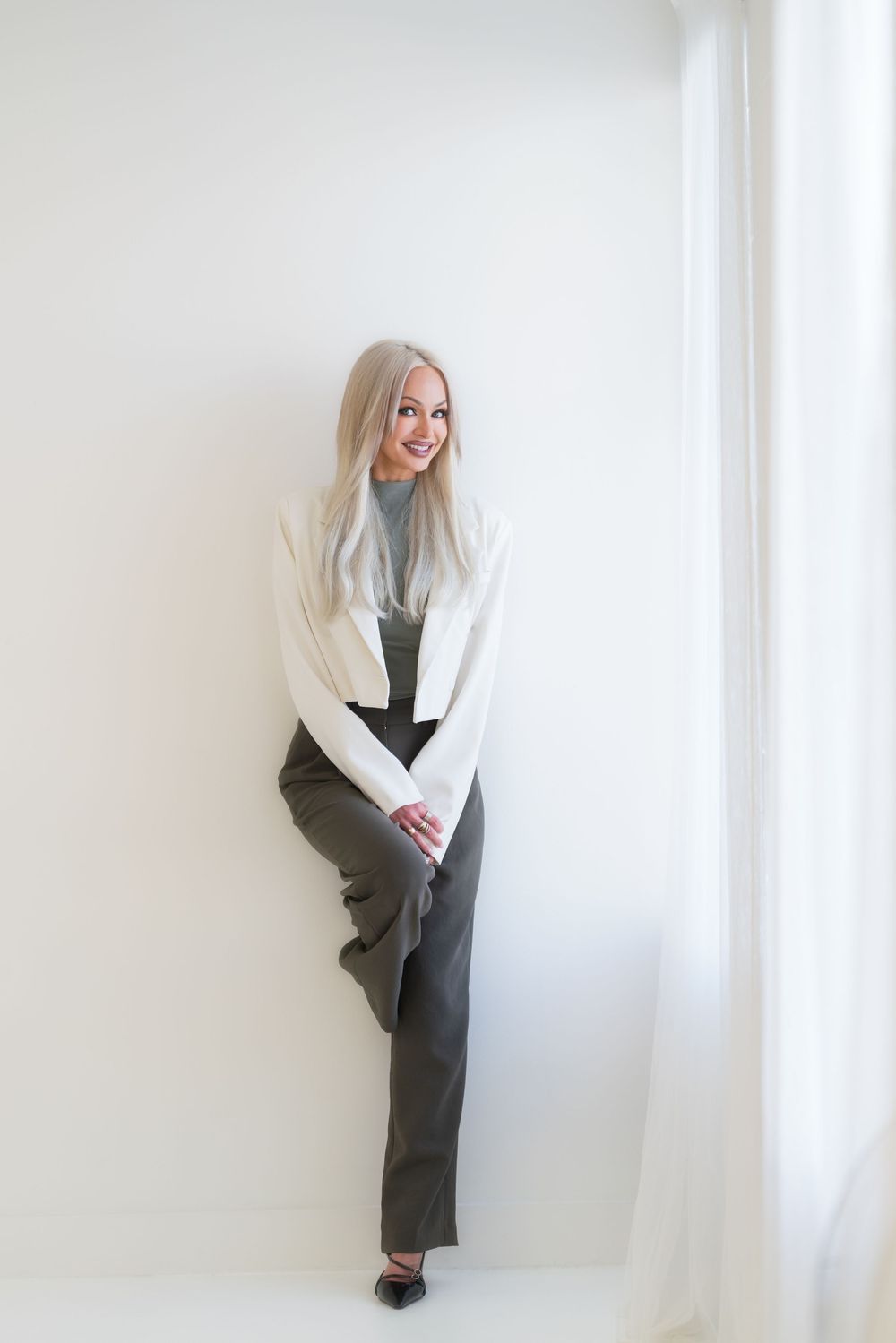 Professional studio portrait of someone wearing a white sweater and gray pants leaning against a white wall.