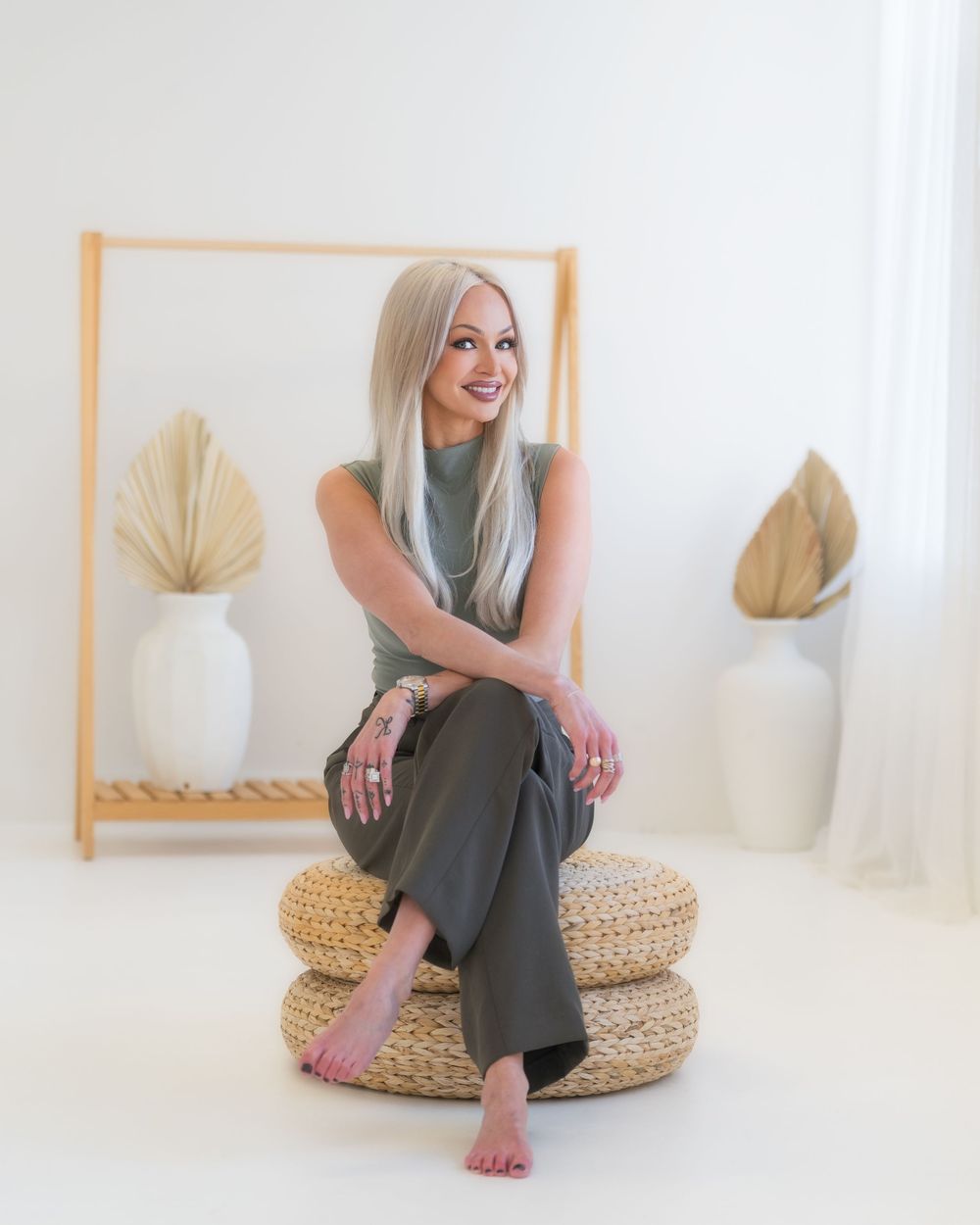 Minimalist white room with someone seated on woven poufs, wearing gray outfit with dried palm leaves in vases.