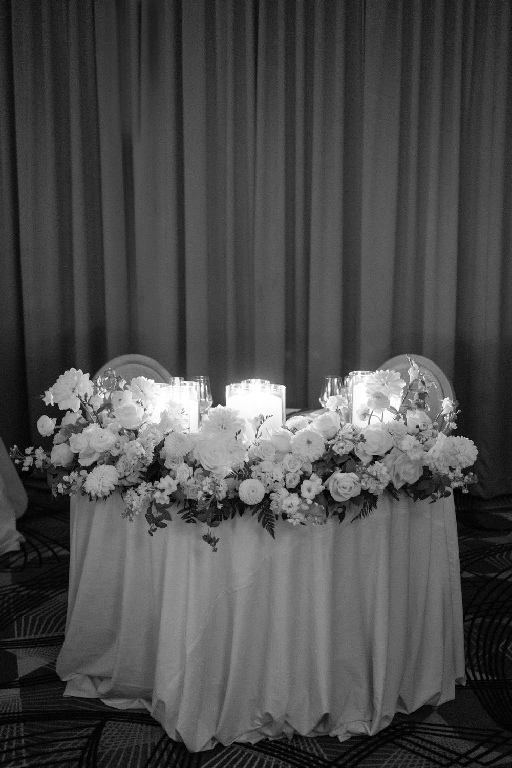 Elegant sweetheart table decorated with white roses, glowing candles and floral arrangements on white linens for wedding reception.