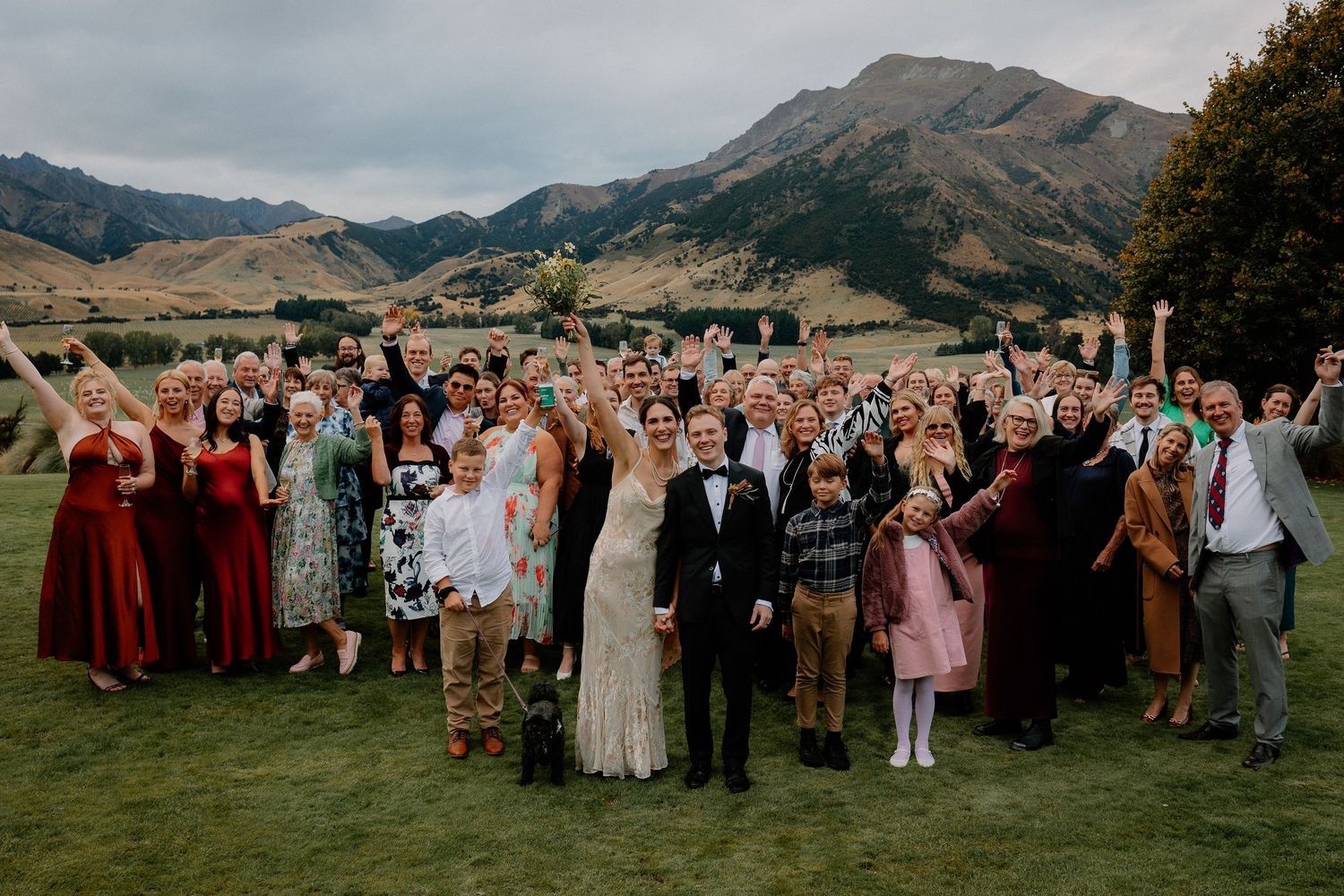 A large wedding party gathers for a group photo on a grassy hill with scenic mountains in the background.