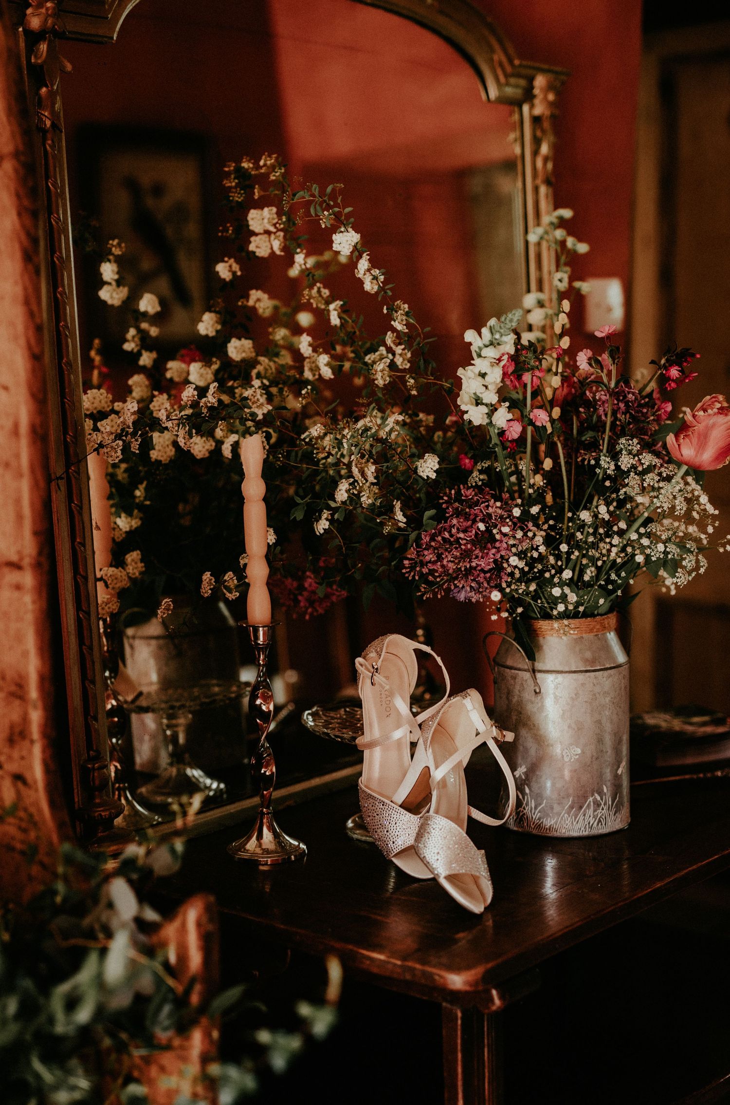 Elegant white strappy wedding shoes next to vintage floral arrangements and mirror on dark wooden vanity table.