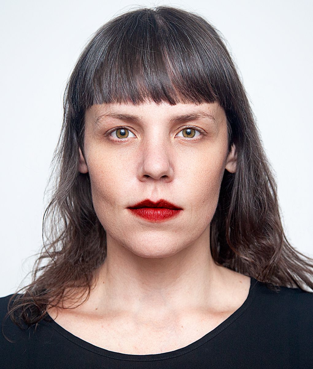 Studio headshot portrait with dark hair, bangs, and bold red lipstick against white background.