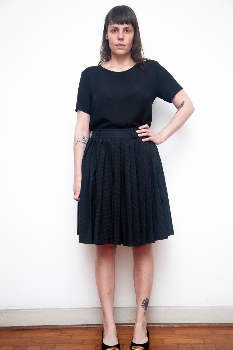 Full length view of someone standing against white wall wearing black t-shirt and pleated knee-length skirt.
