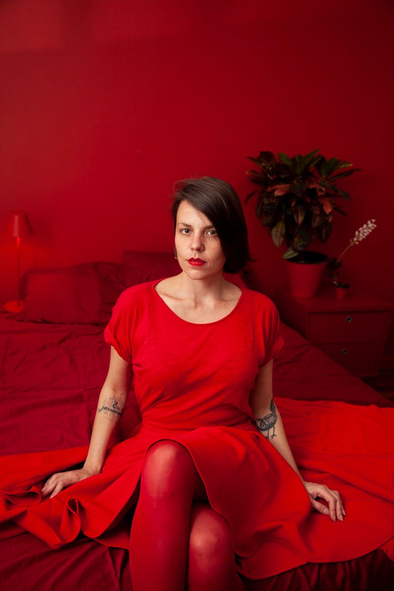 Woman in red dress sits on red bedding with matching red wall and potted plant in monochromatic scene.