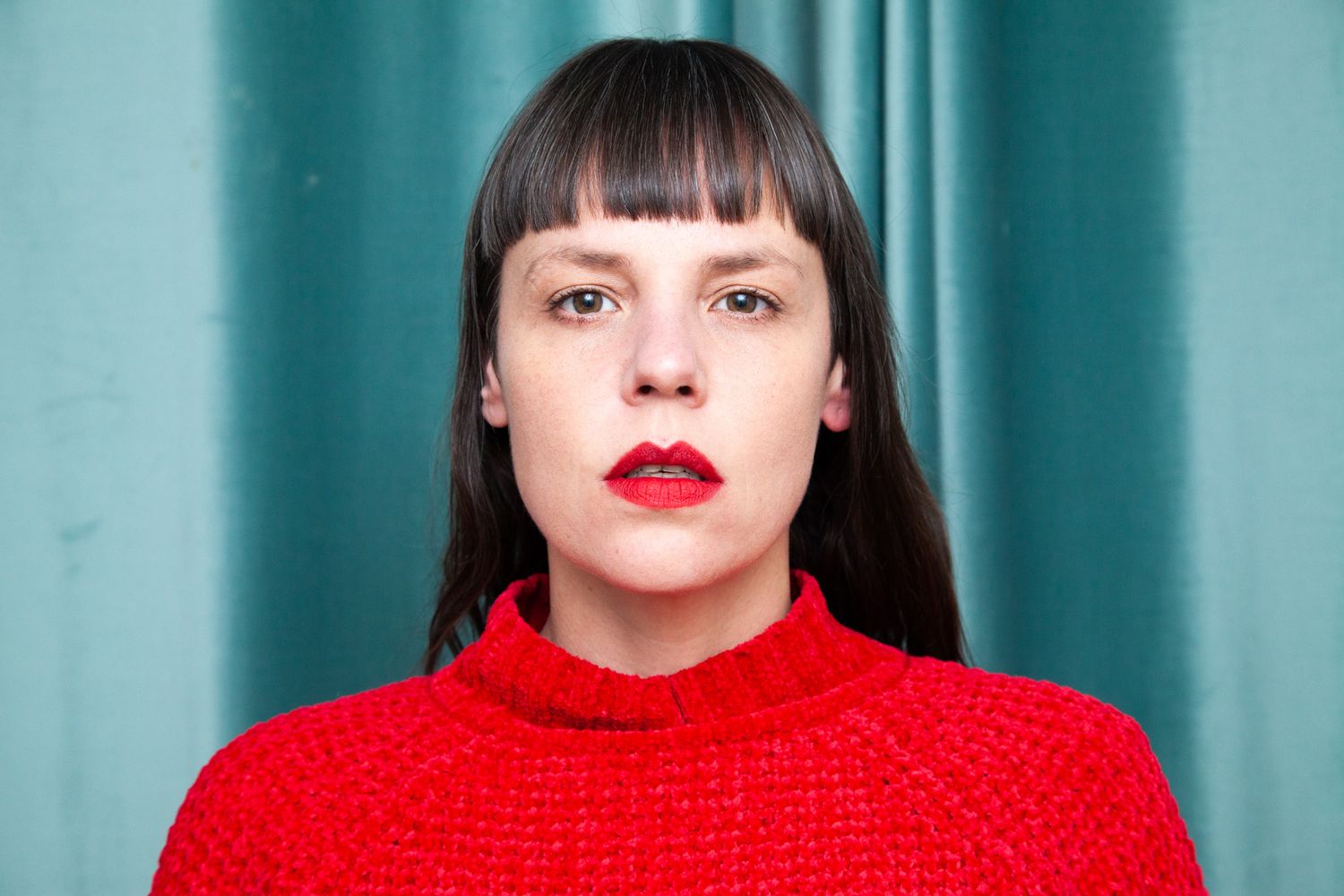 Close-up portrait against a turquoise wall wearing a bright red sweater and bold red lipstick.