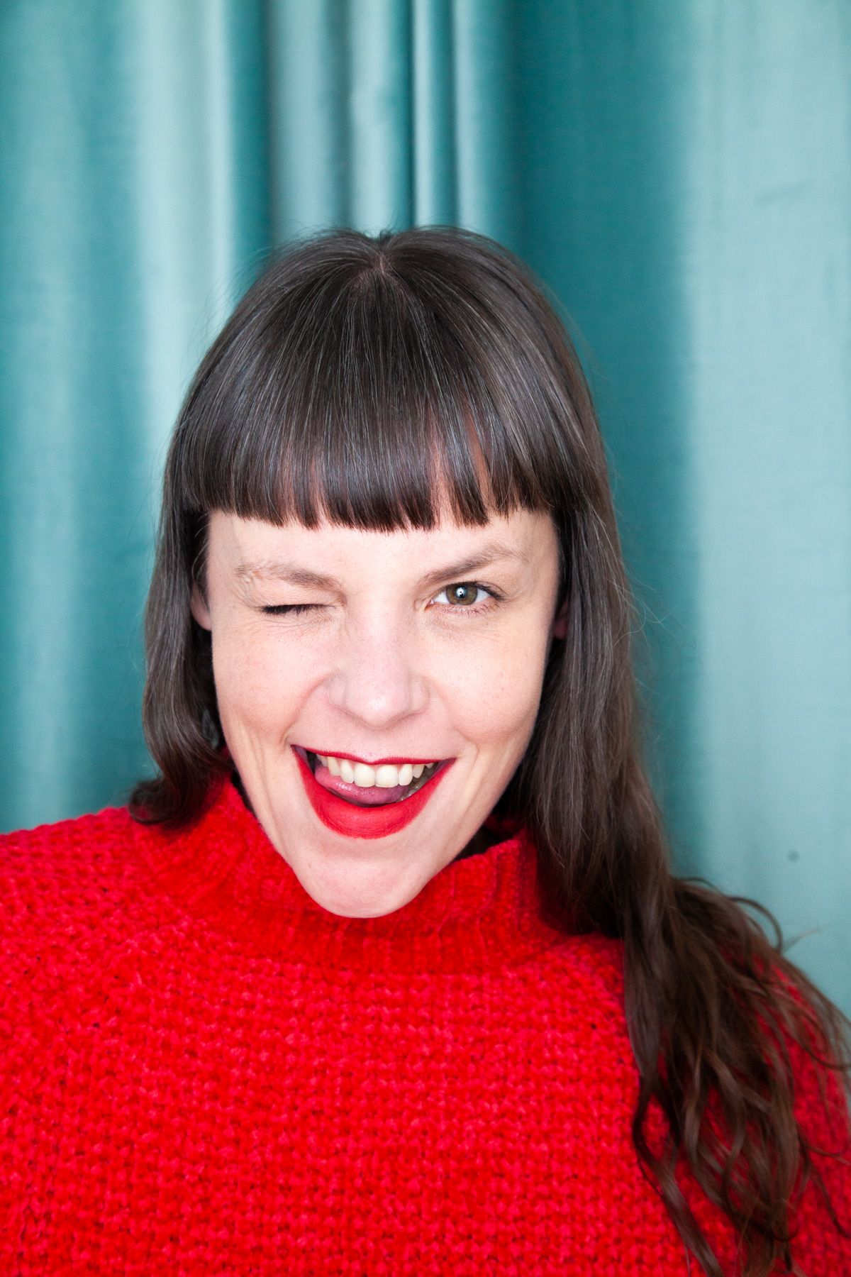 Person with dark bangs and long hair wearing a bright red sweater smiles joyfully against turquoise curtain background.