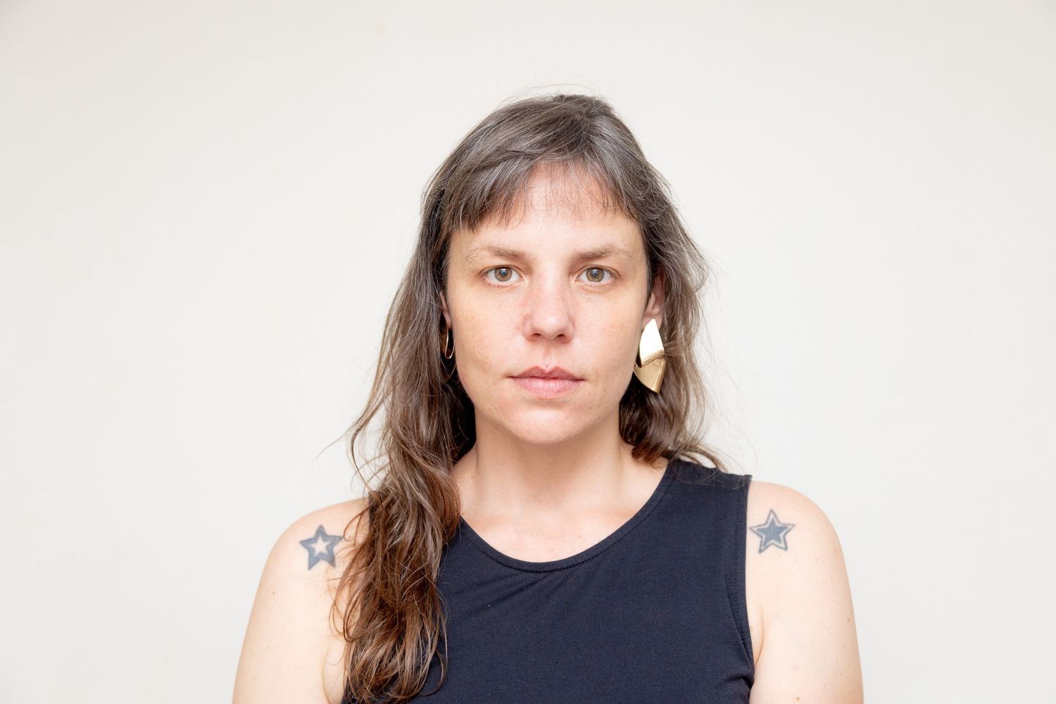 Person with long brown hair and visible tattoos wearing a black sleeveless top against a plain light background.
