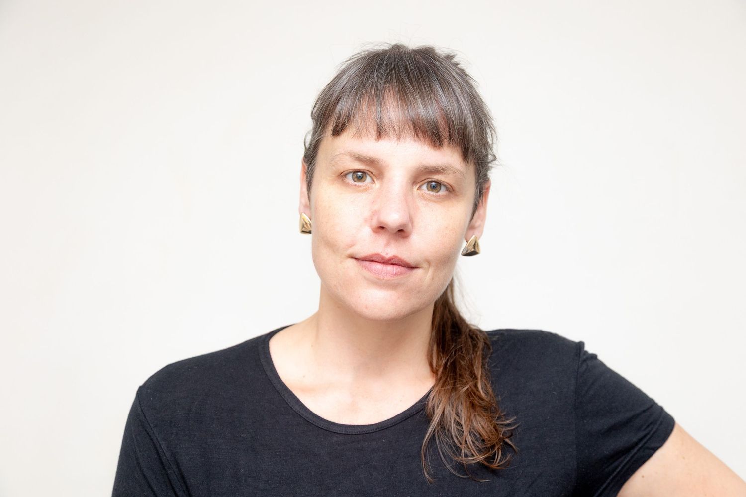Portrait shot of someone with long brown hair in a ponytail wearing a black t-shirt against a white background.