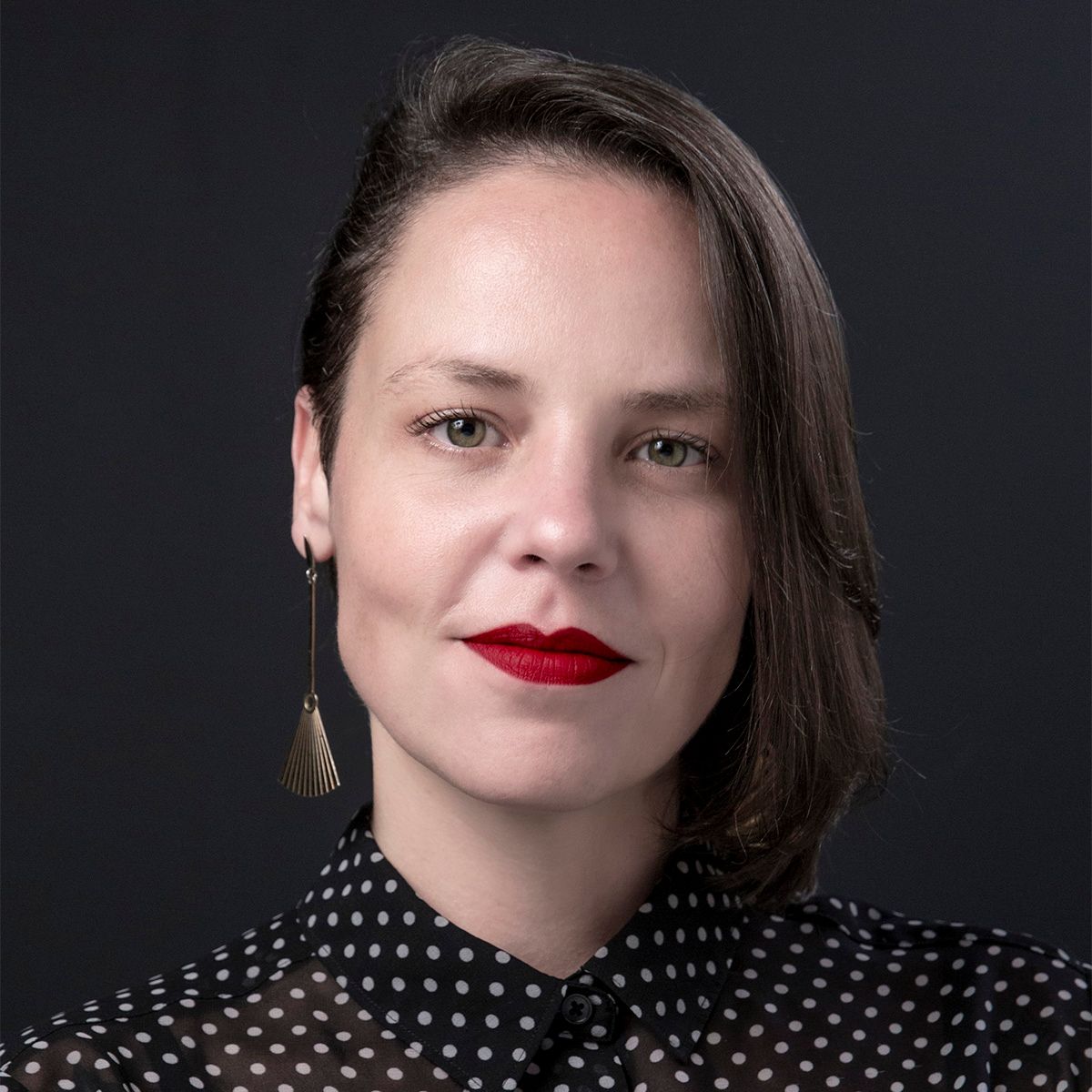 Professional portrait of a person with red lipstick wearing a polka dot blouse against dark background.