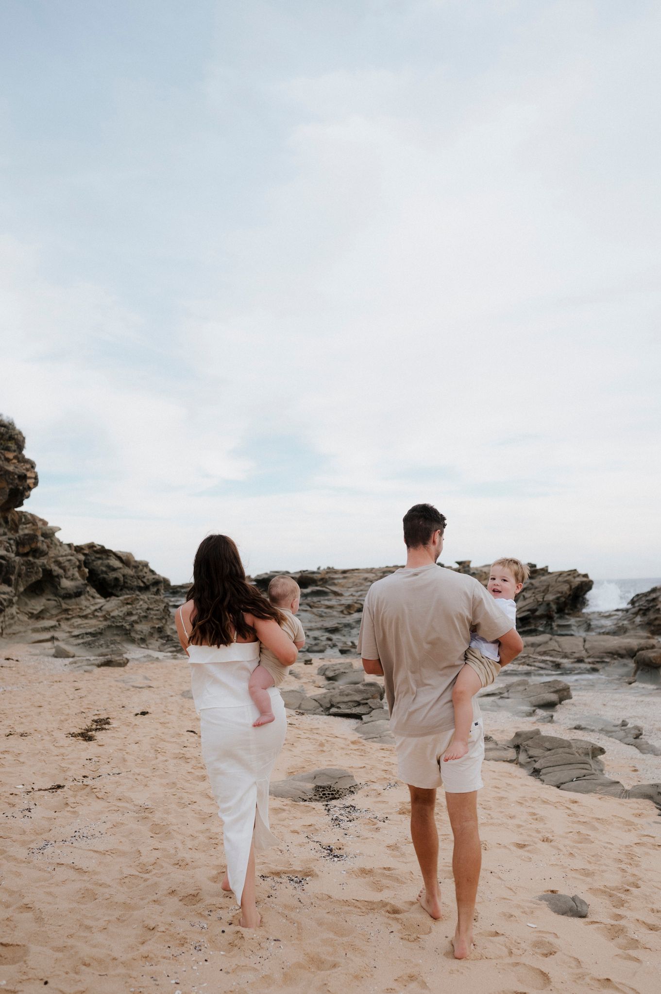 Family walking barefoot on rocky beach shore at sunset.