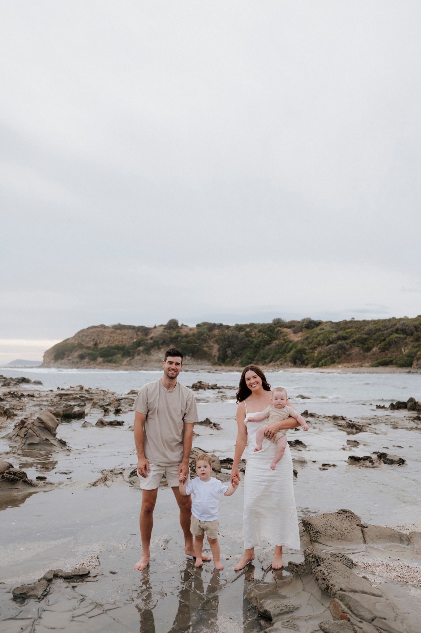 Family of four standing barefoot on rocky beach during cloudy day.