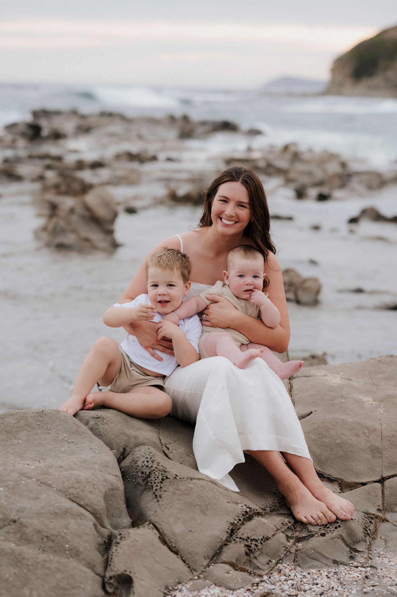 Mother and children sitting on coastal rocks with ocean view background.