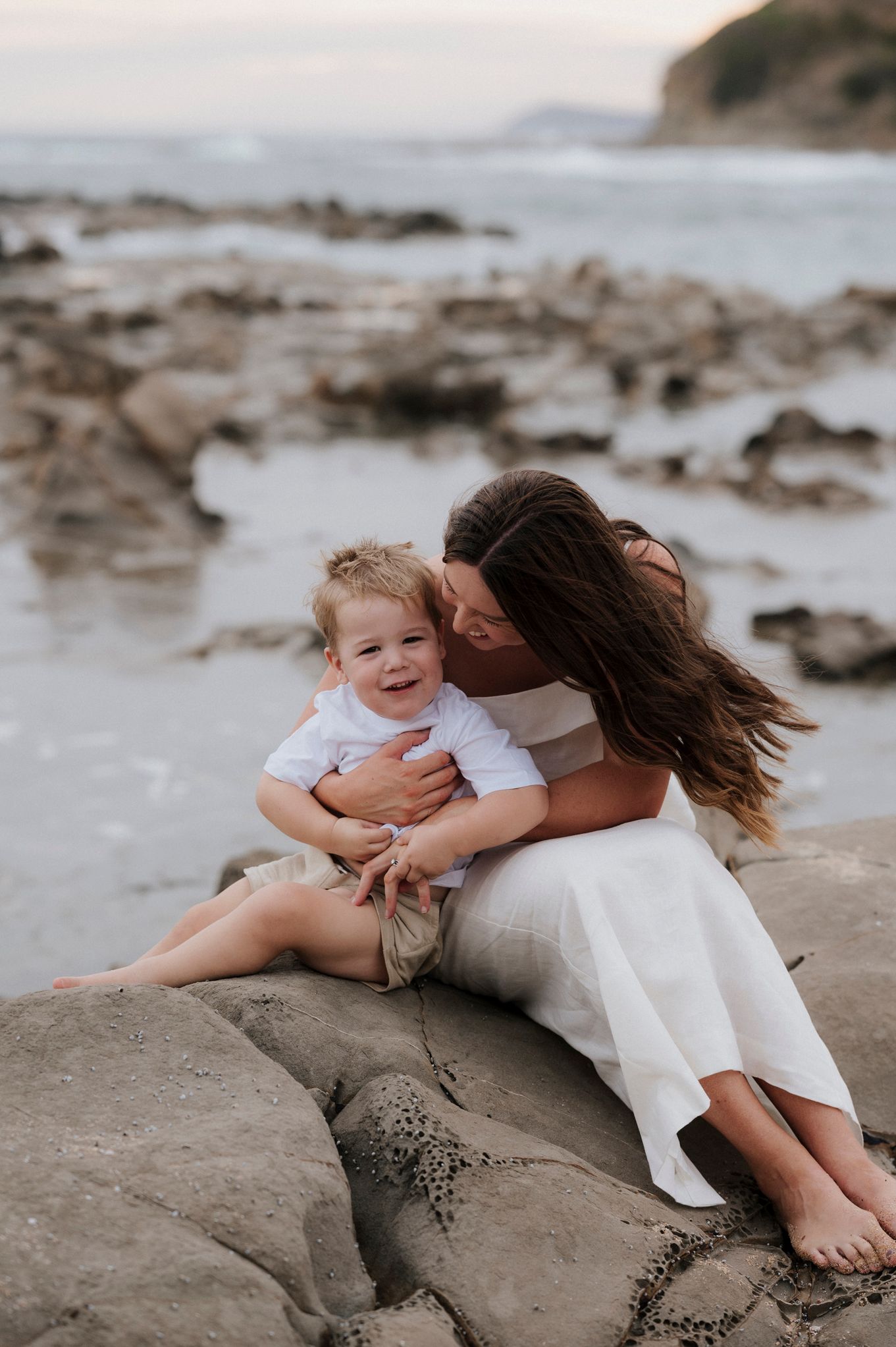 A joyful moment shared between parent and child sitting on coastal rocks at sunset.
