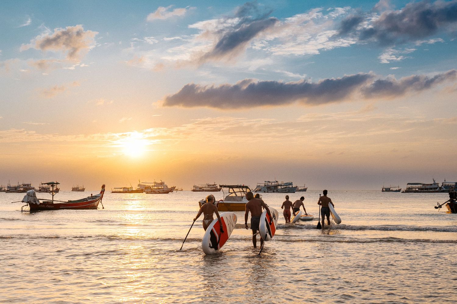 Silhouetted fishermen wade in shallow waters at sunset with traditional longtail boats anchored in the background.