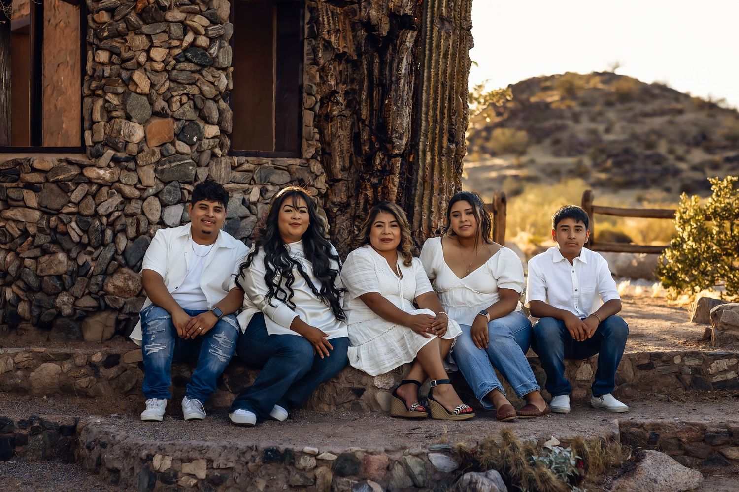 A group of people in white tops and jeans sit together on a rustic stone wall with mountains in the background.