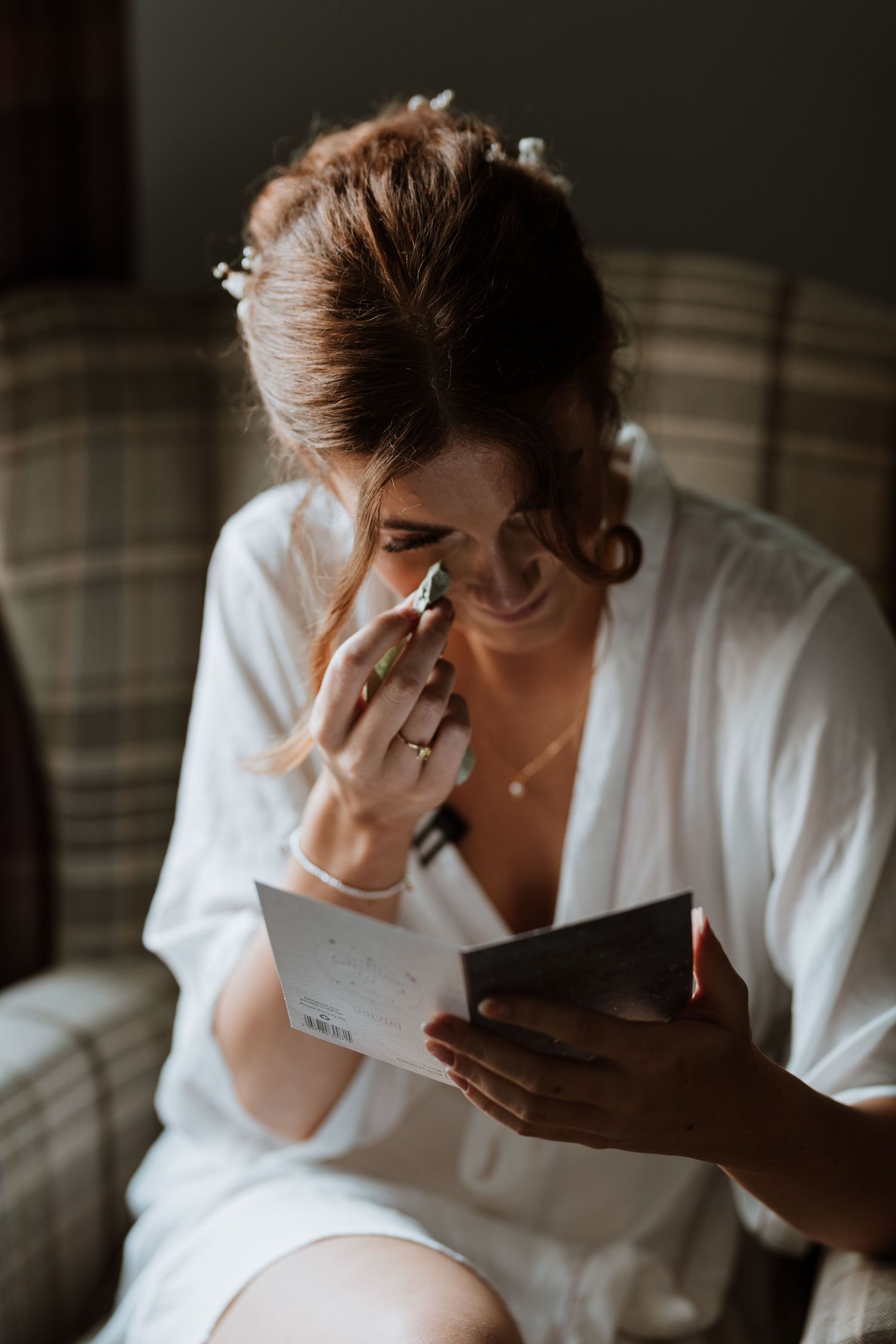Bride in a white robe wipes tears while reading a letter on a plaid chair at Stanford Farm.