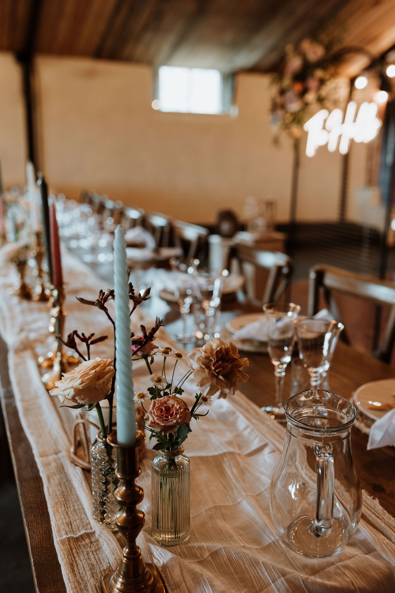 Rustic wedding reception table setting with light blue taper candles, pink flowers in glass vases, and crystal glassware.