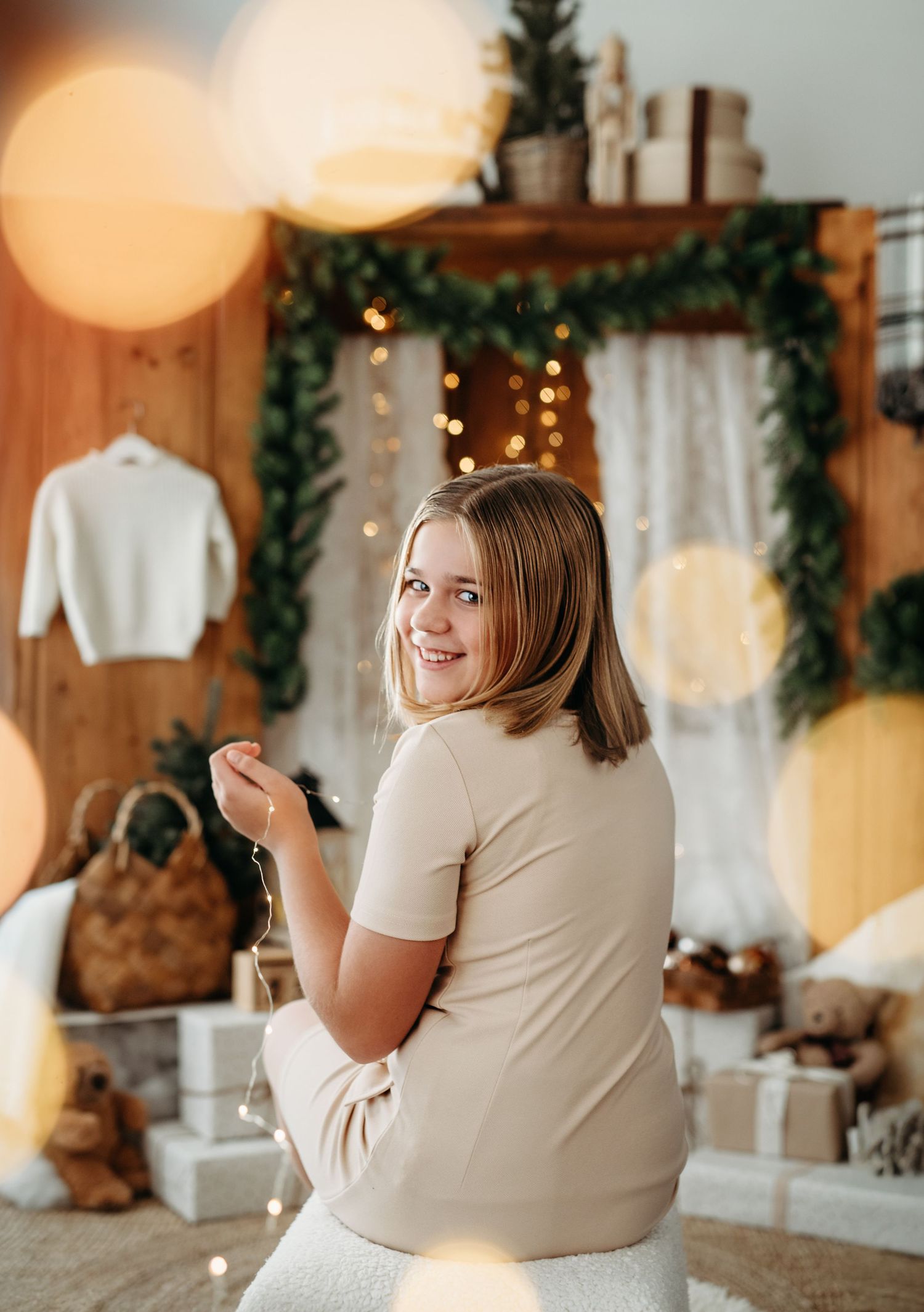 A holiday scene with someone in cream clothing against twinkling lights and greenery decorations.