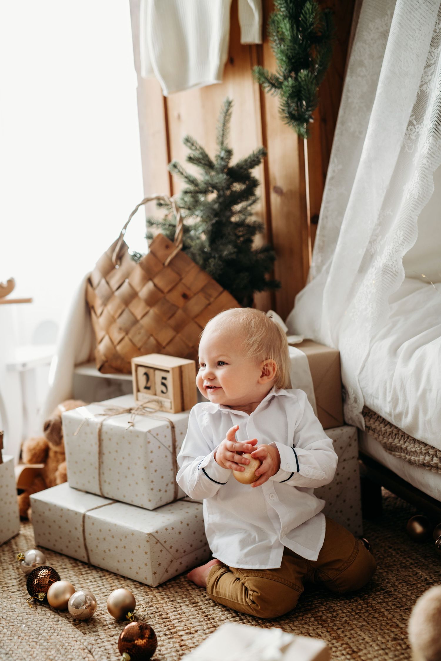 Baby in white outfit sits among Christmas decorations and presents with pine tree branches and ornaments in background.