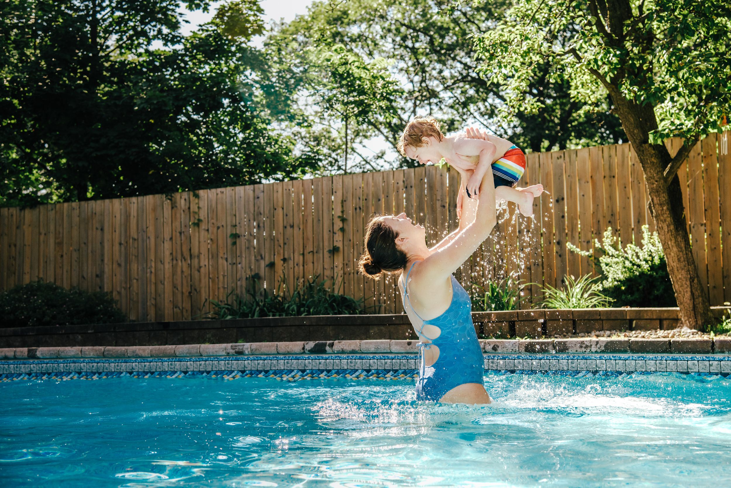 A person in a blue swimsuit playfully lifts a toddler high above them in a swimming pool on a sunny summer day.
