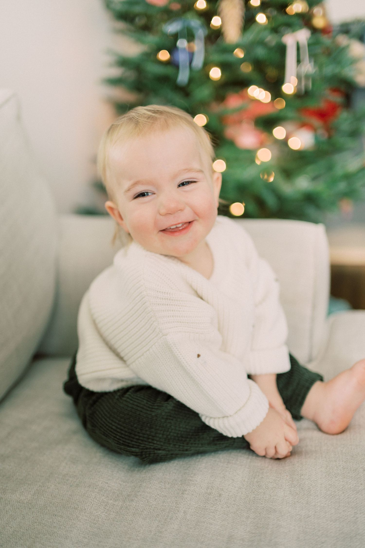 Baby in white sweater smiles while sitting on gray couch near Christmas tree.