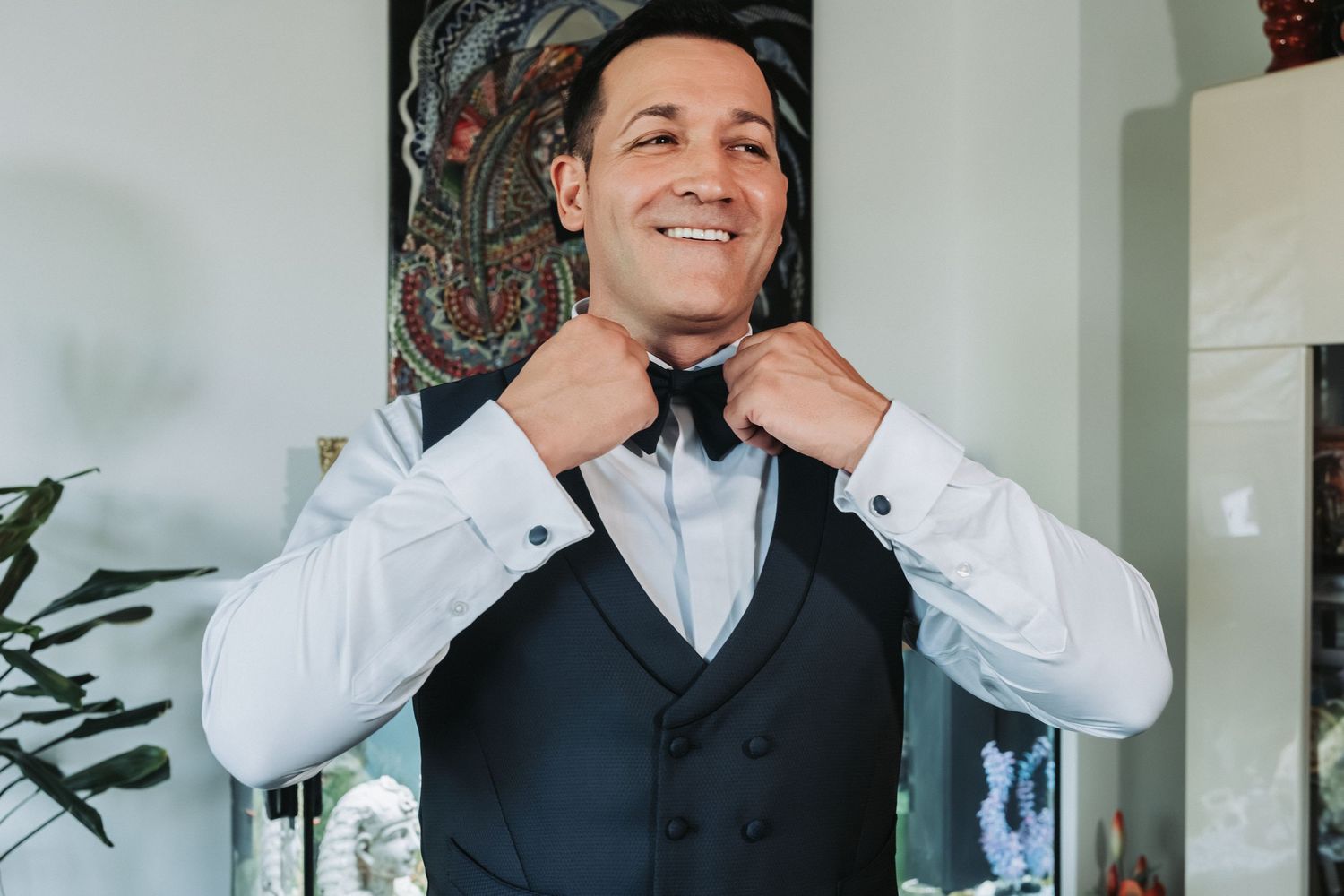 Groom adjusts his bowtie while getting ready in a room with artistic wall decor.