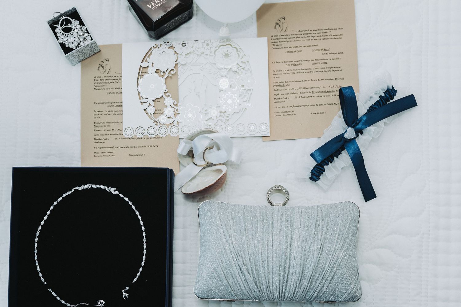 Wedding accessories including invitation, jewelry, clutch and decorative items arranged on white surface.
