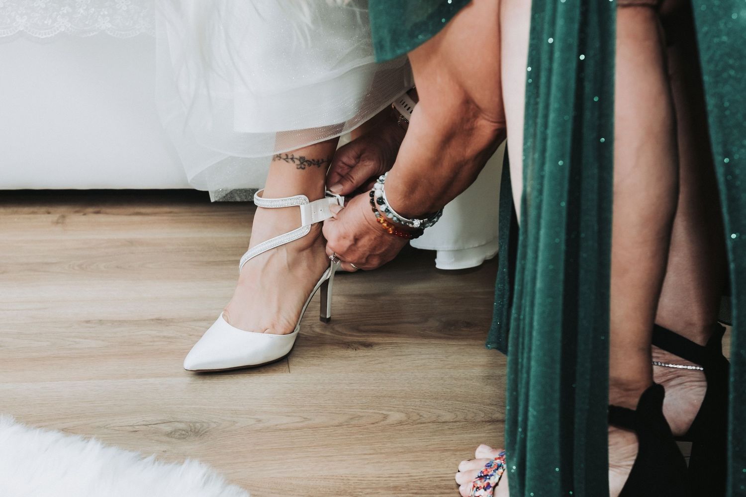 Someone fastening an elegant white bridal shoe on a hardwood floor with green curtains in the background.