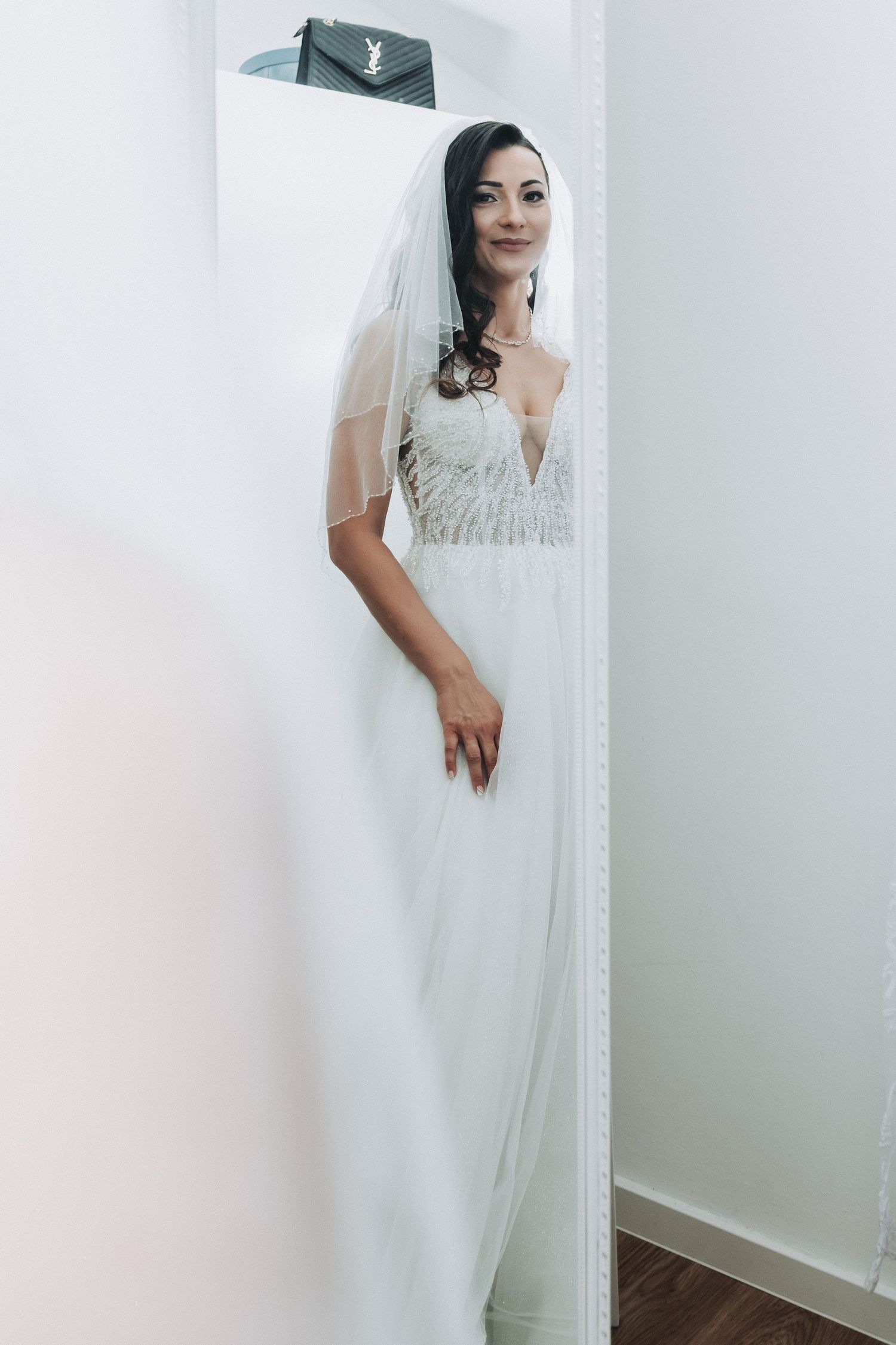 An elegant bride in a white lace wedding gown and veil poses against a light wall.