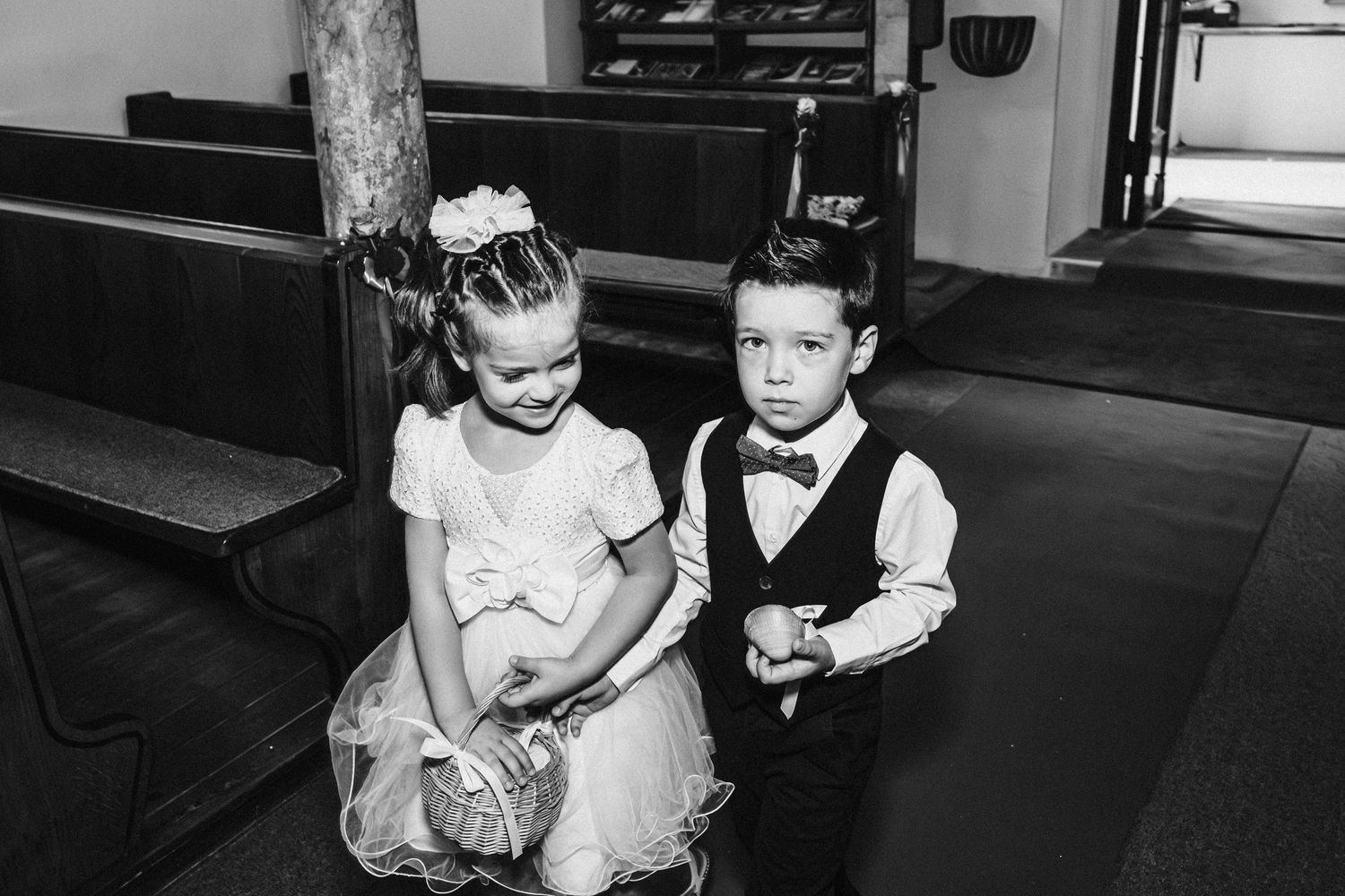 A black and white photo of two young children in formal attire sitting on a church pew.