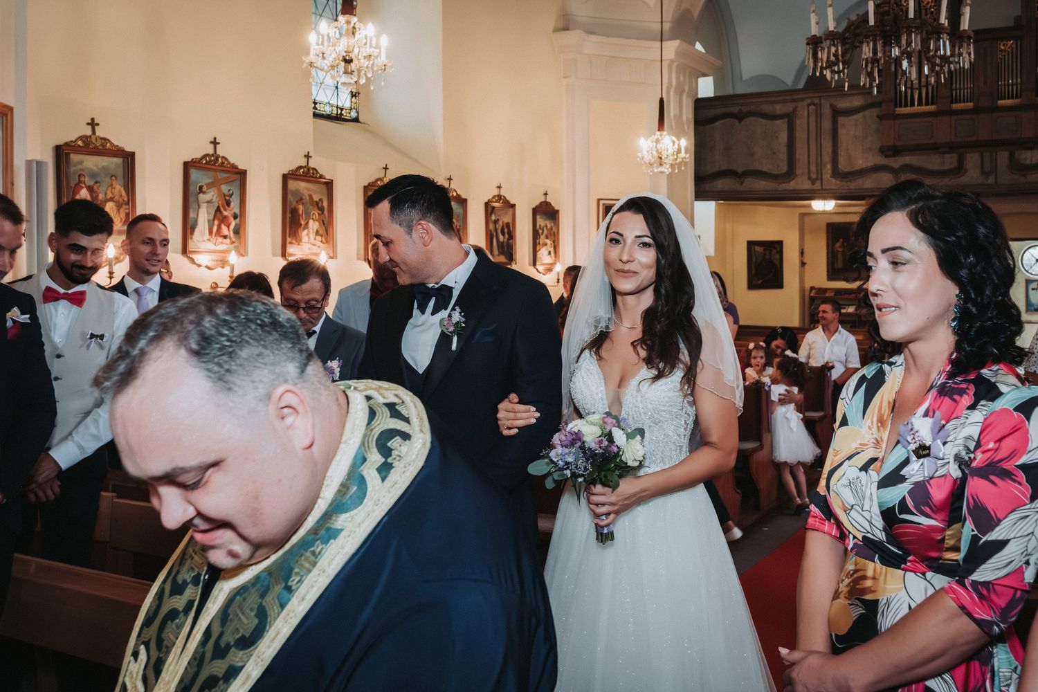 Wedding ceremony in progress inside a traditional church with ornate decorations and hanging chandeliers.