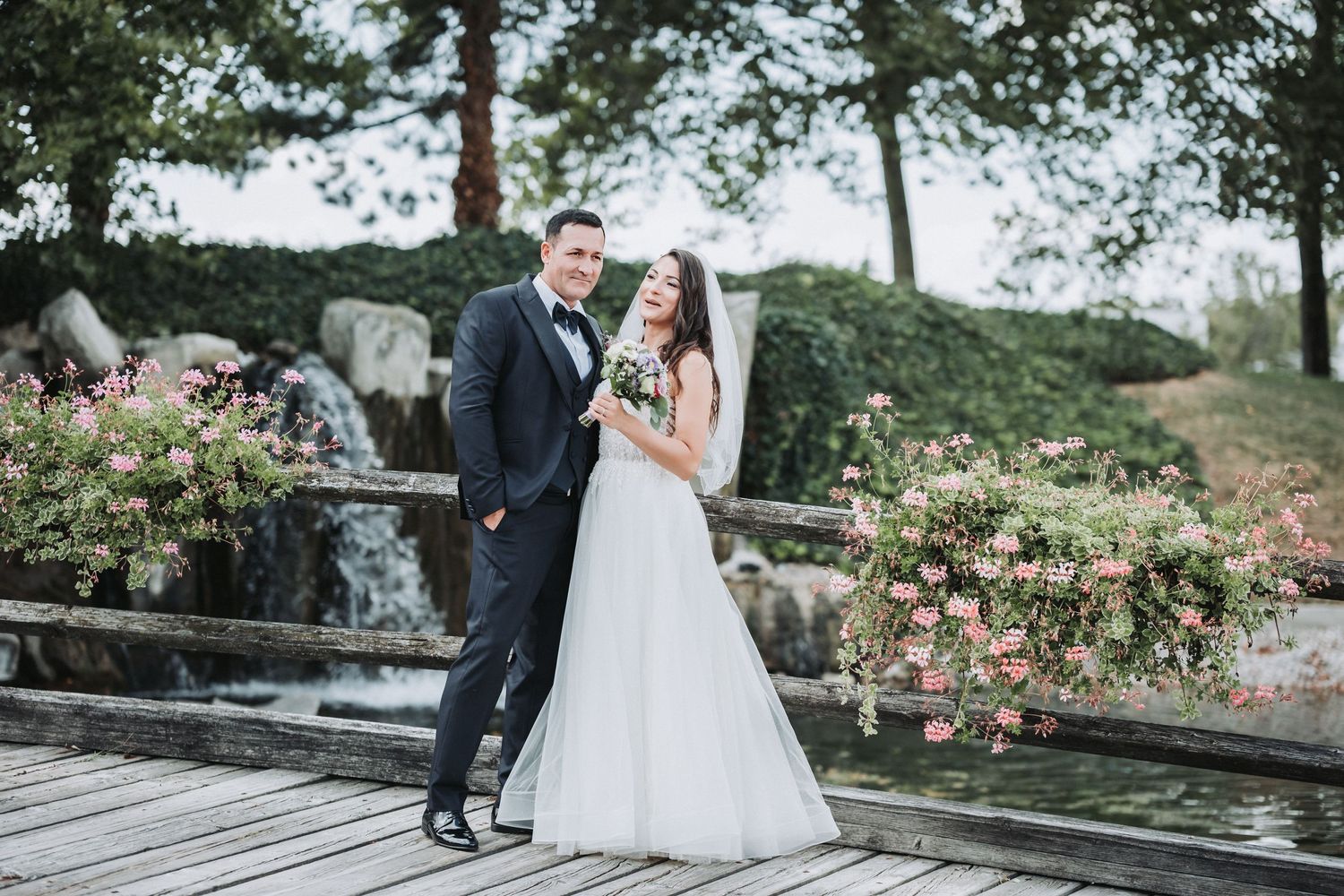 Bride and groom pose on wooden deck with blooming flowers in background.