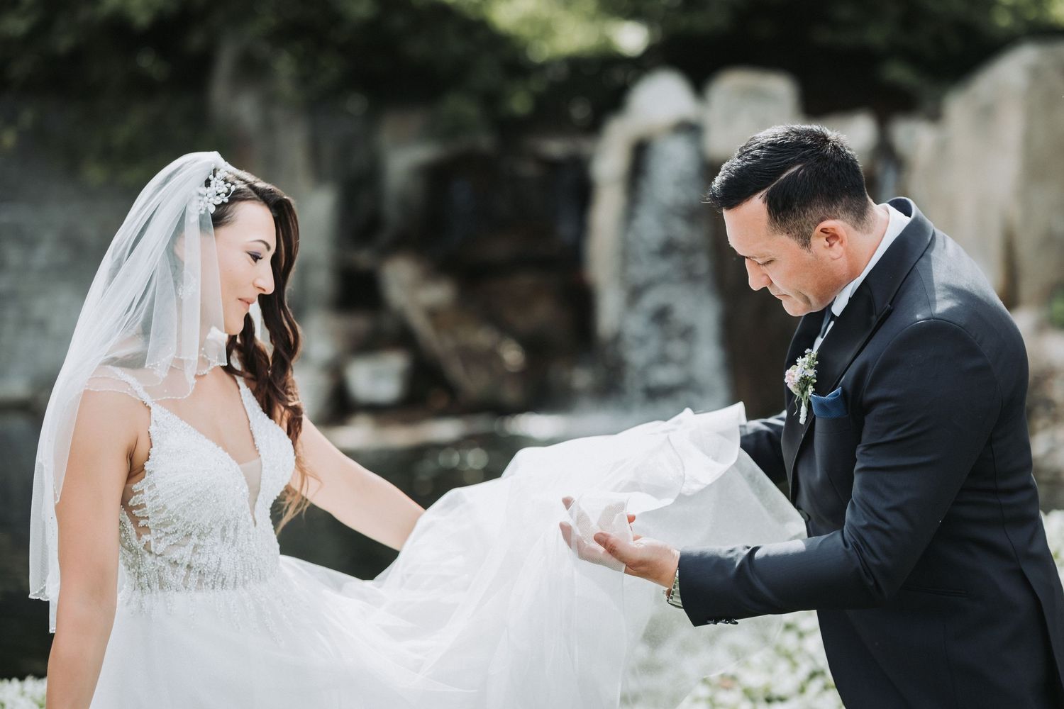 A couple in wedding attire share an intimate moment outdoors as the groom delicately holds the bride's veil.