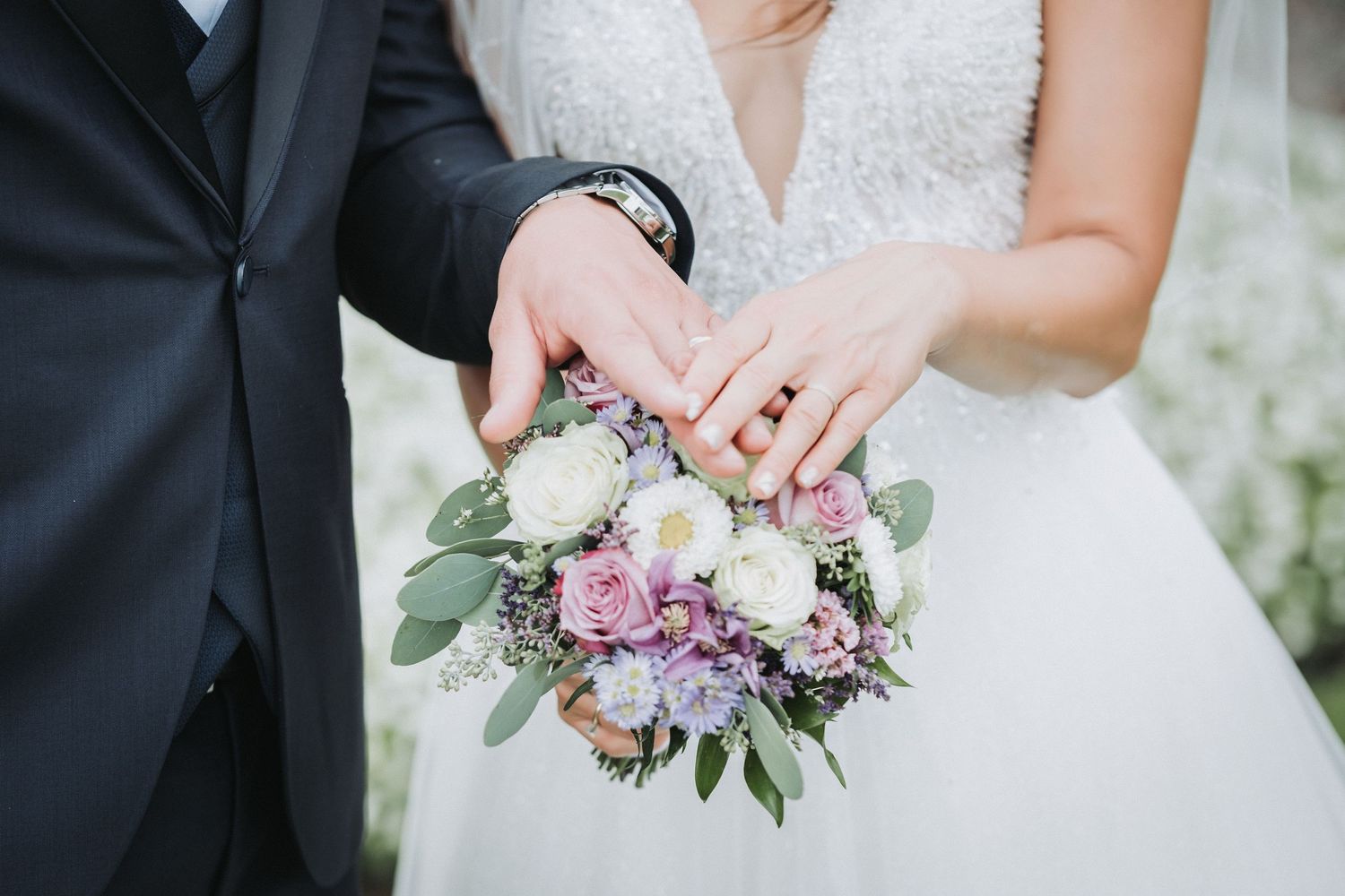 Close-up of newlyweds' hands holding a beautiful bouquet with pink and purple flowers and greenery.