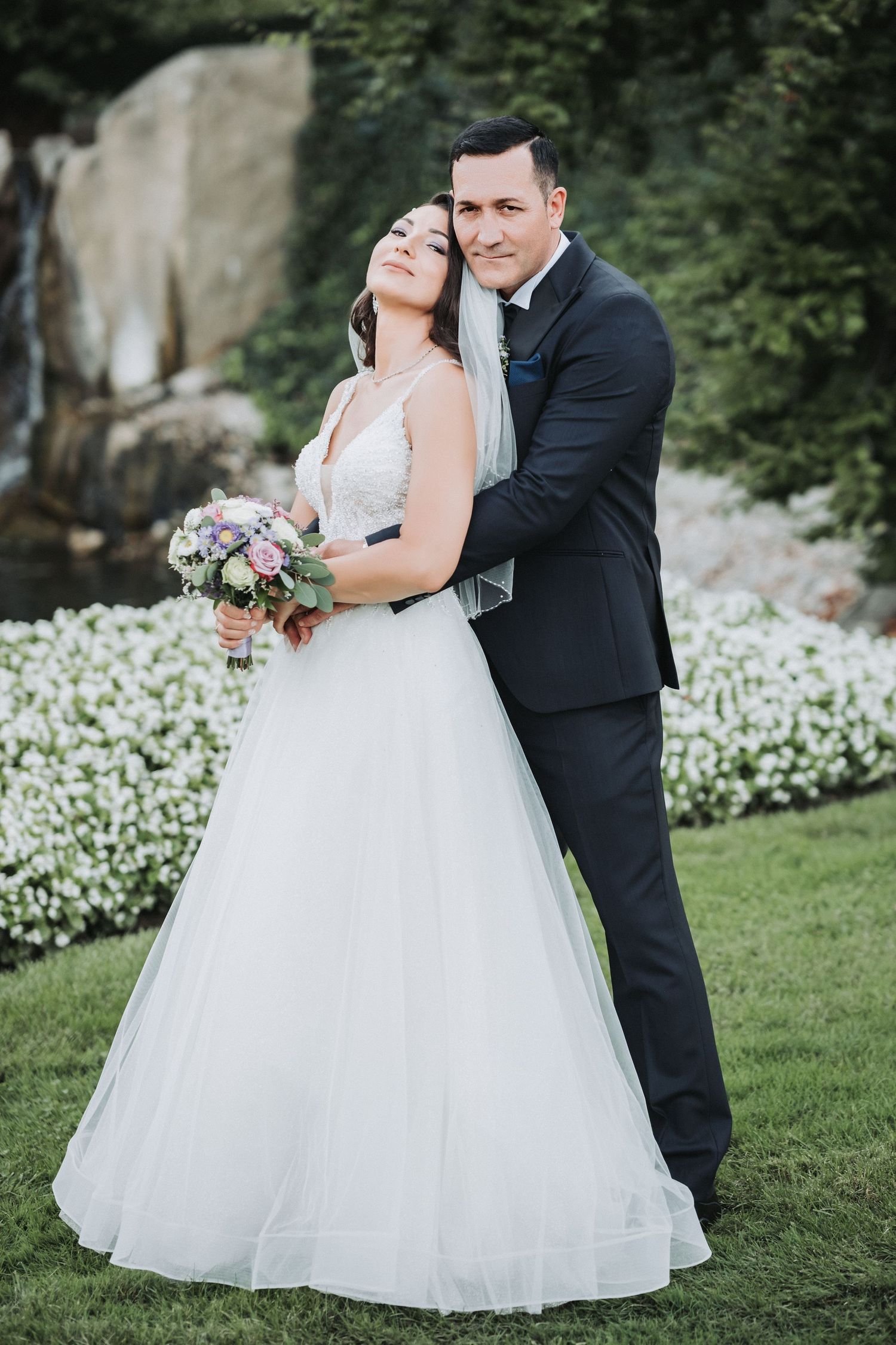 Newlyweds embrace in a garden setting with white flowers blooming in the background.