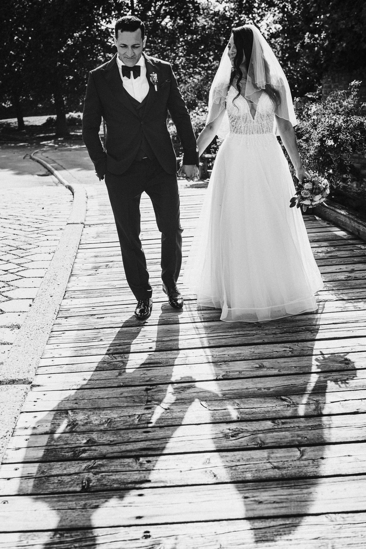 Black and white photo of newlyweds walking on a wooden pathway casting long shadows.