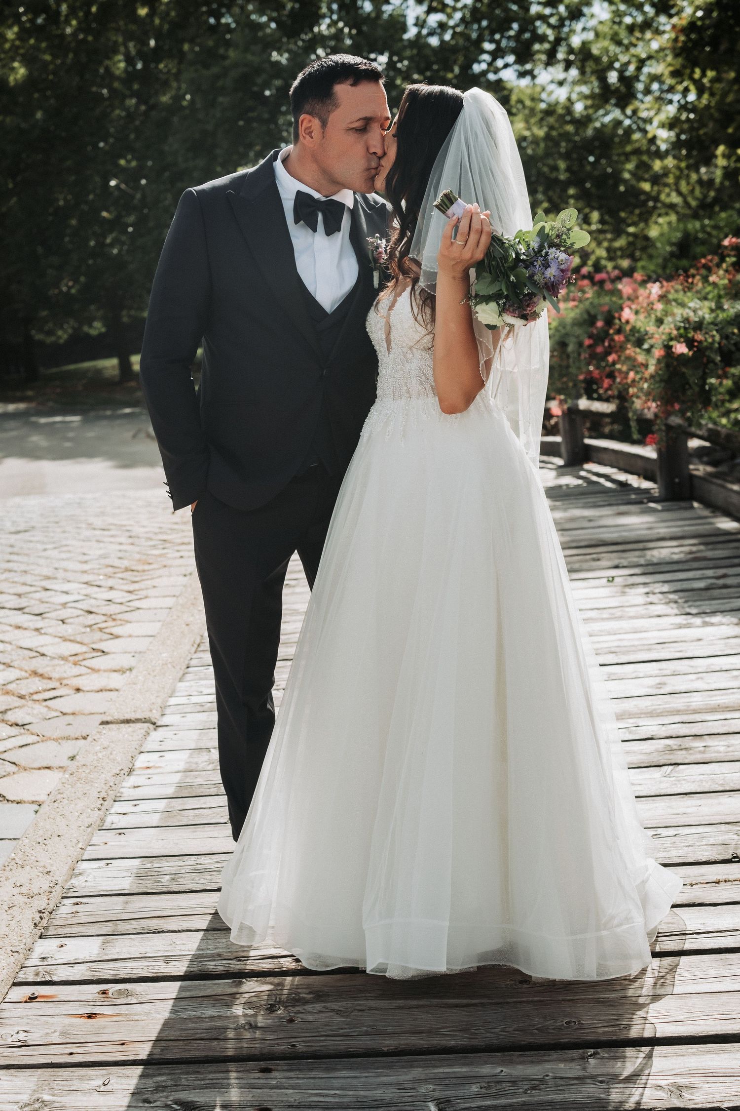 Elegant couple in wedding attire sharing an intimate moment on a sunlit wooden boardwalk.
