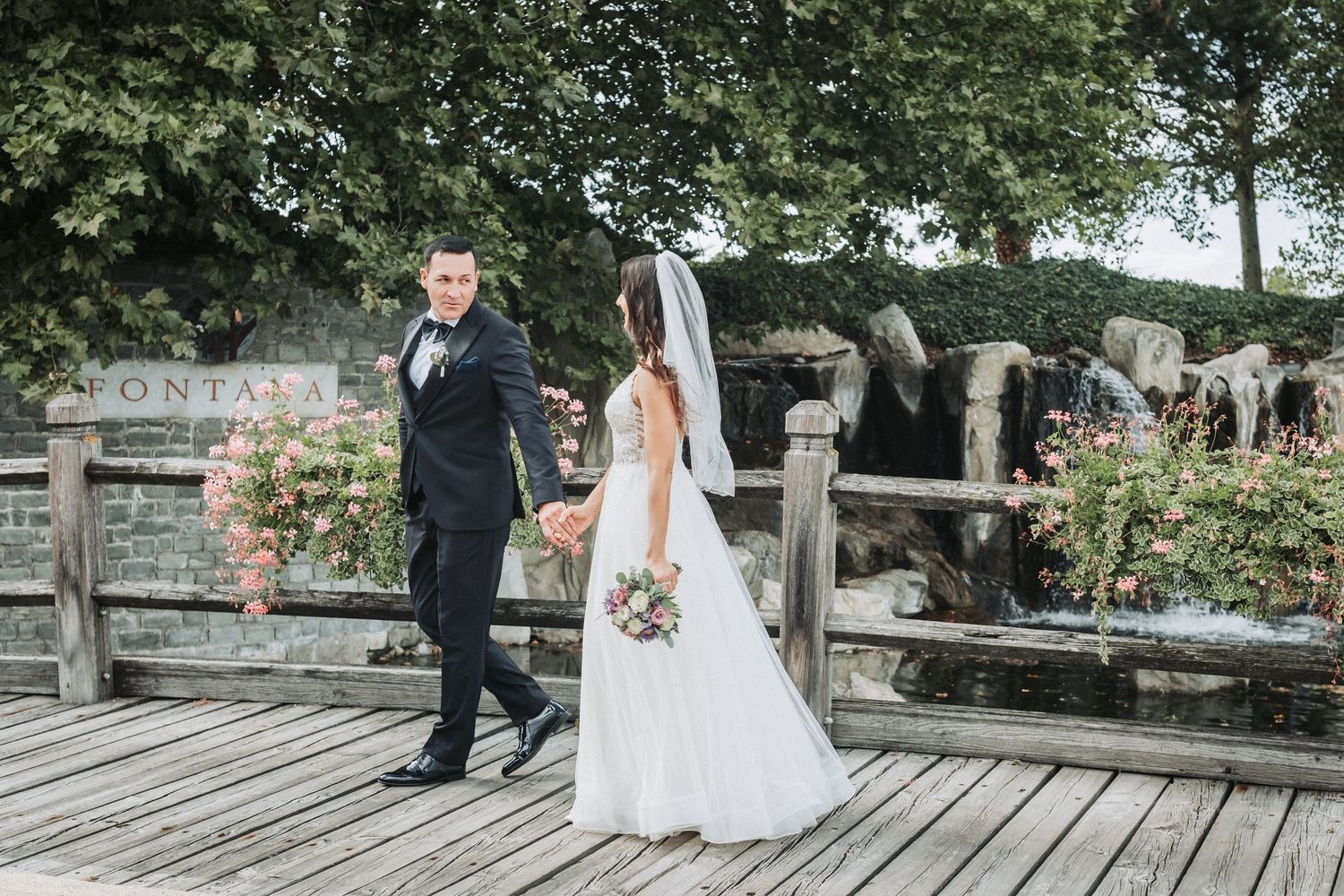 Newlyweds in formal attire standing on wooden deck surrounded by greenery.