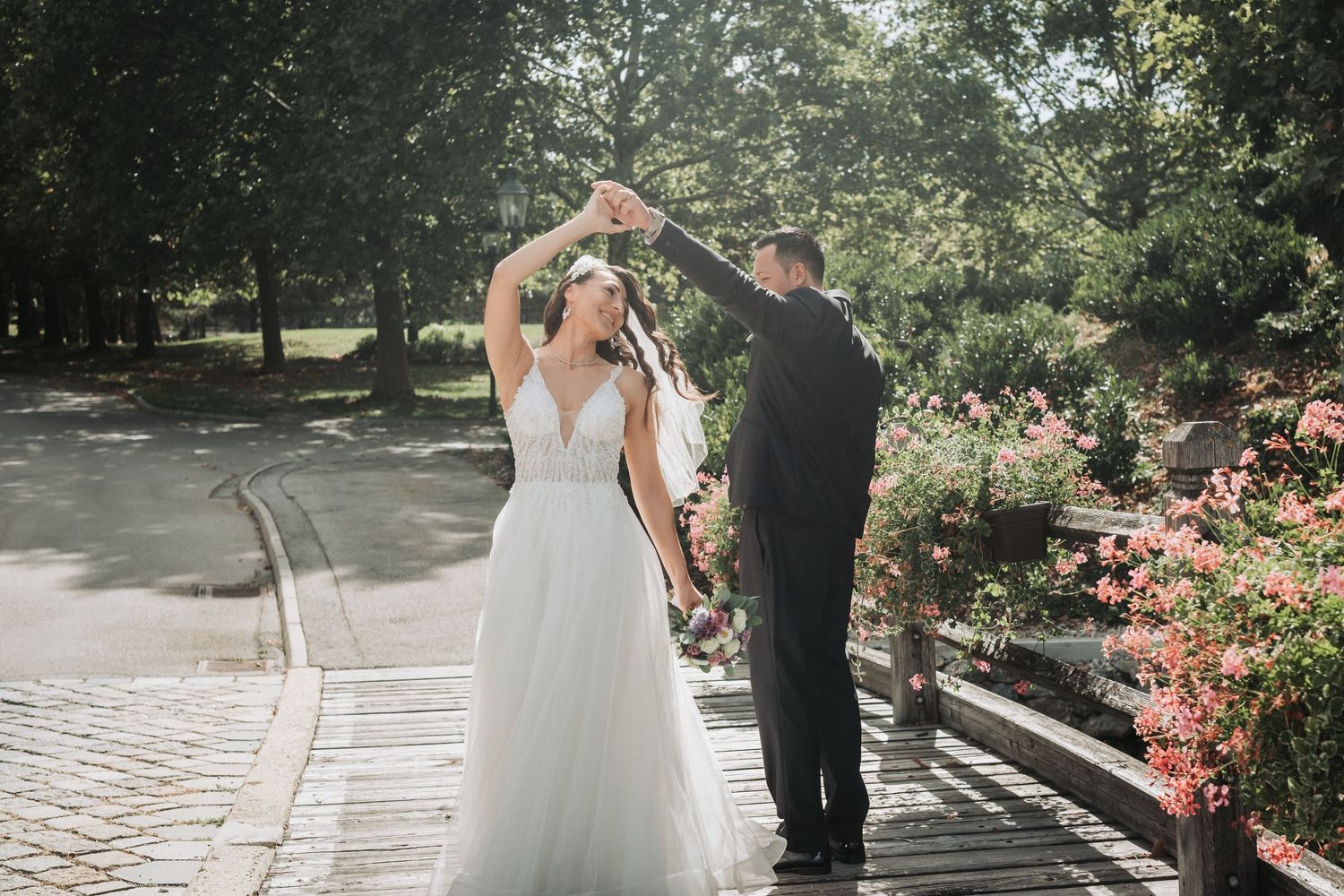 Couple dancing together on a sunlit pathway surrounded by greenery and pink flowers.