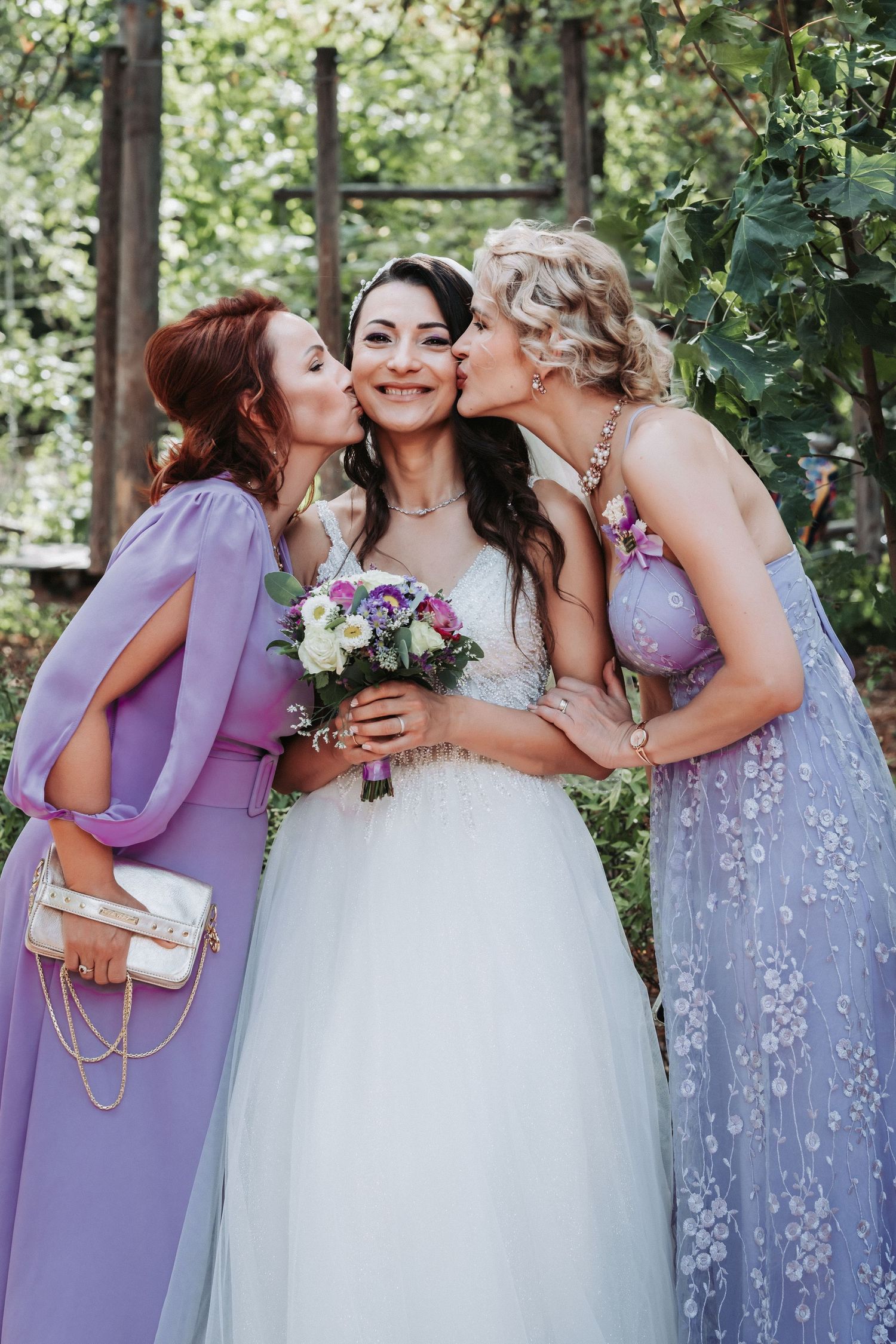 Bride in white dress receiving affectionate kisses while holding purple bouquet.