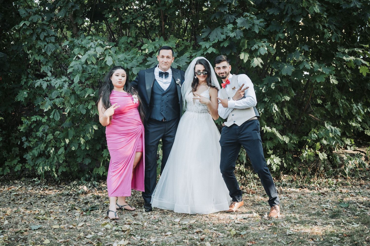 Small wedding group in formal attire posing casually against green foliage.