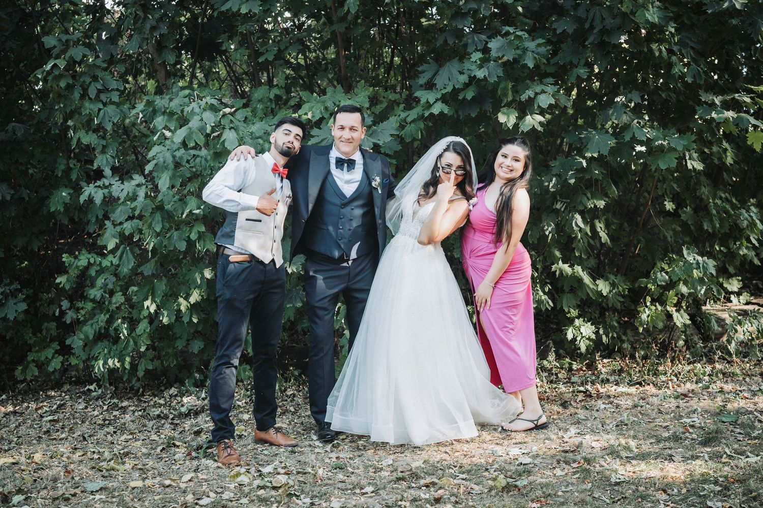 A wedding party poses playfully against a green leafy backdrop outdoors.