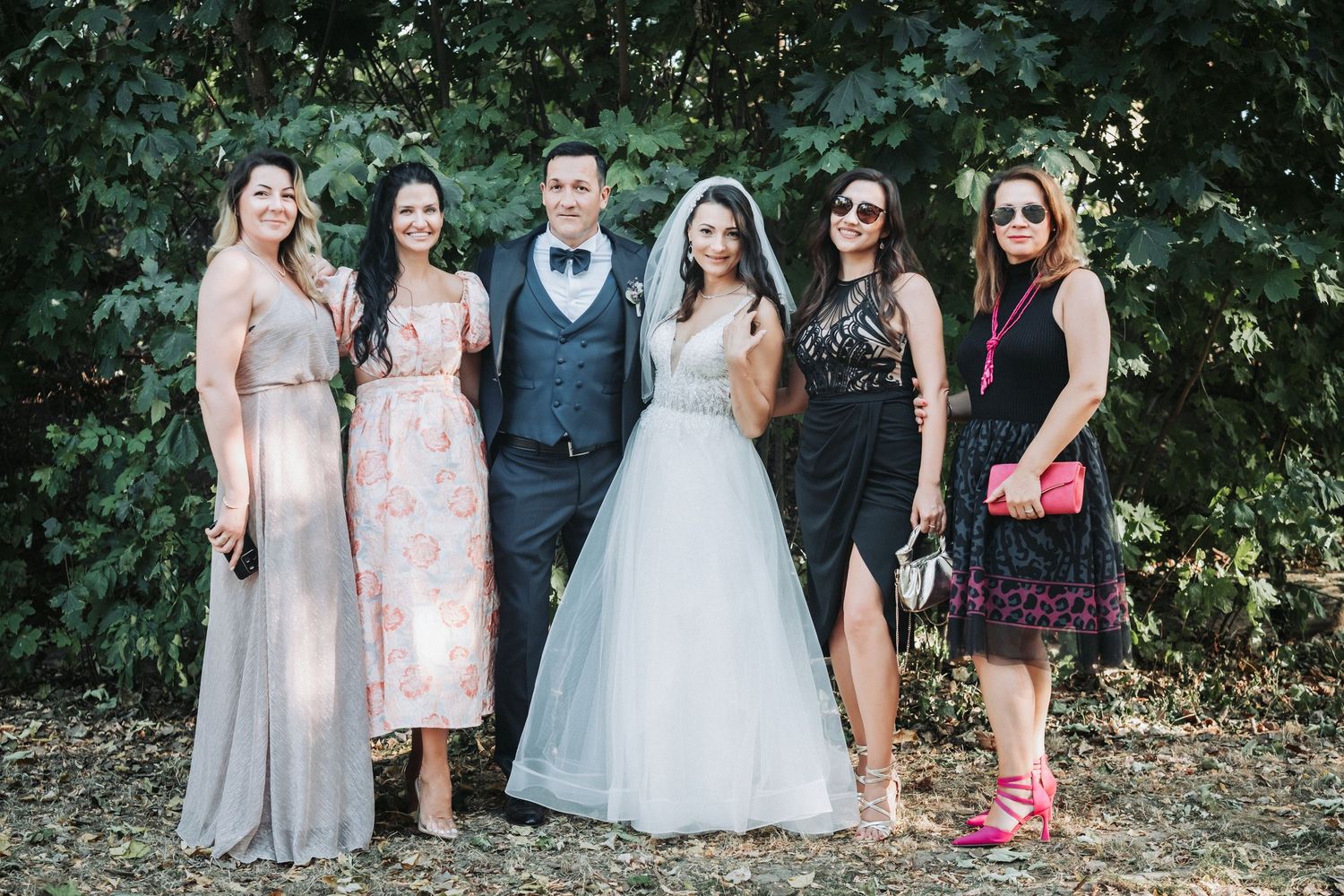Wedding party members in formal attire pose against foliage at an outdoor celebration.