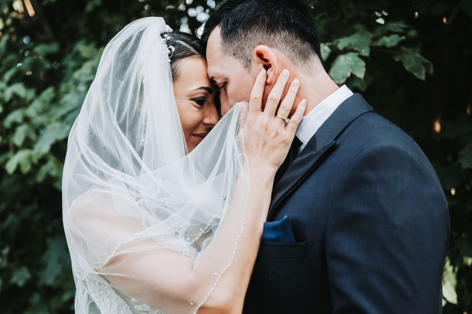 A romantic wedding moment captured as a couple shares an intimate moment with the bride in her veil against greenery.