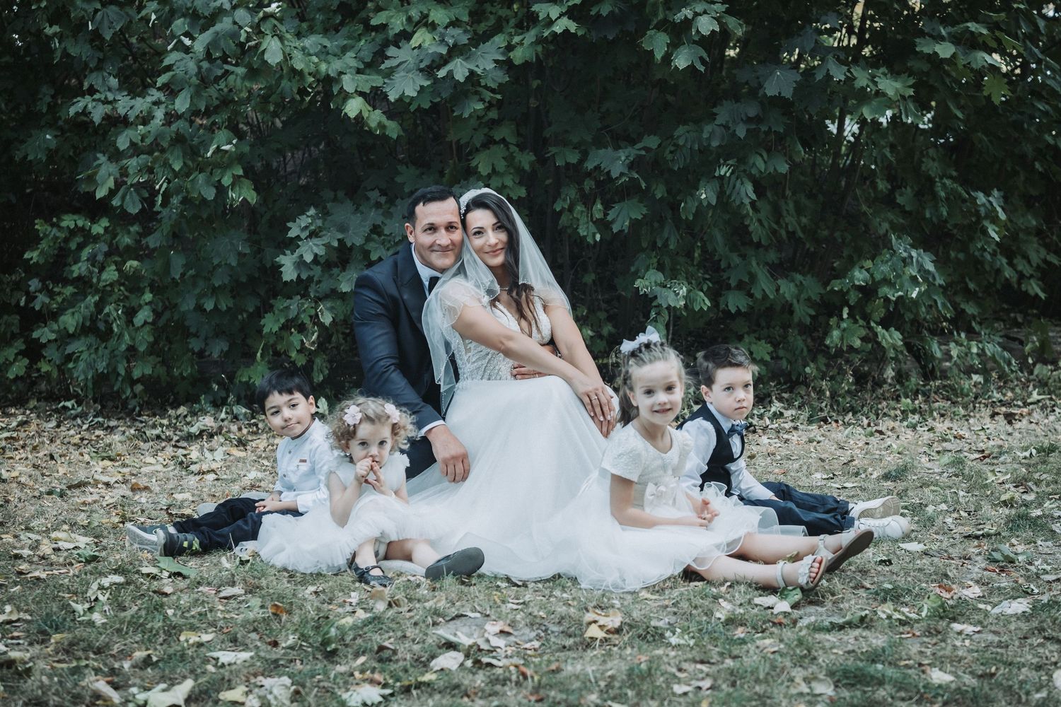 A wedding couple and young children pose on the ground for a formal portrait outdoors.