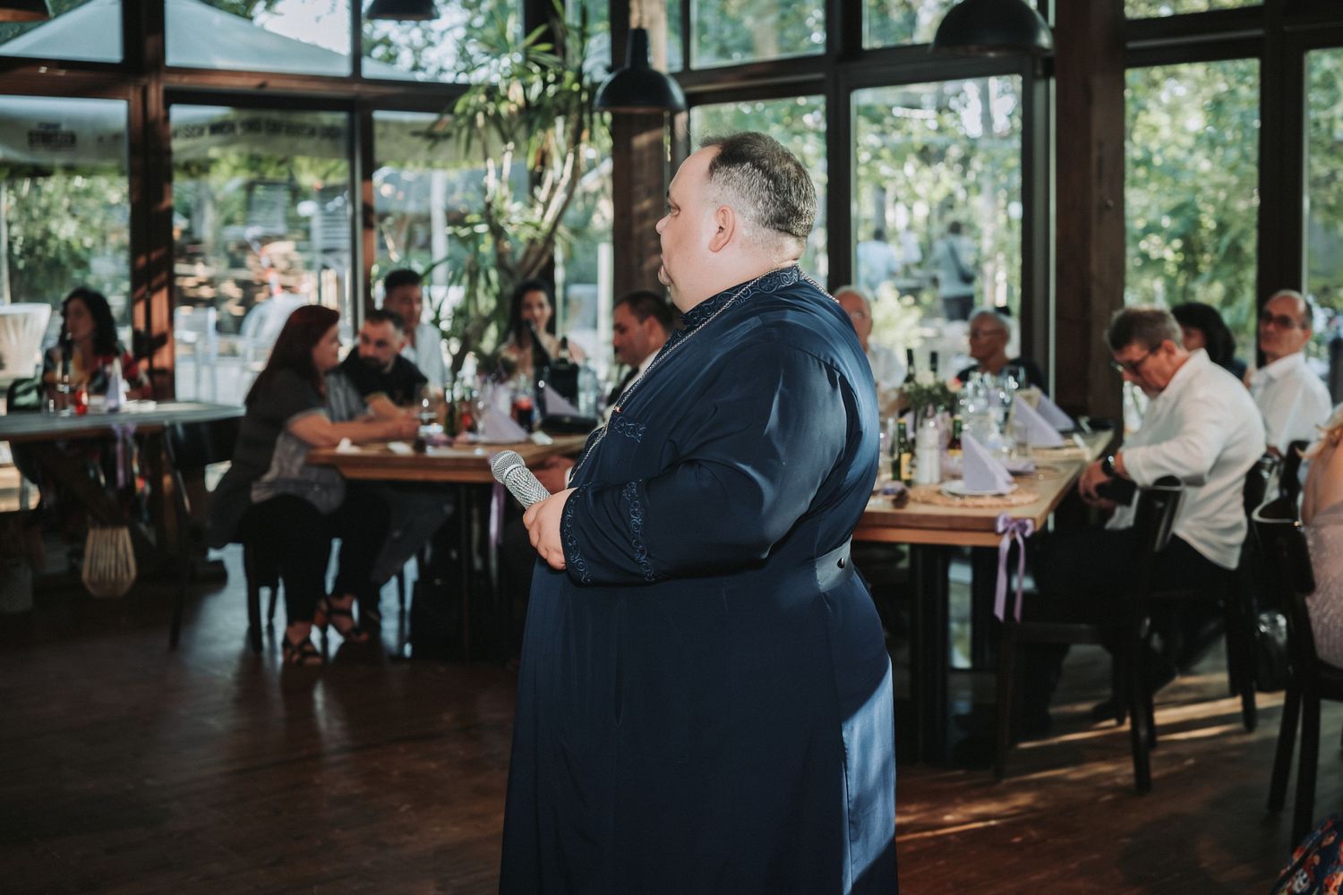 A person in a navy suit gives a speech in a restaurant setting with large windows.