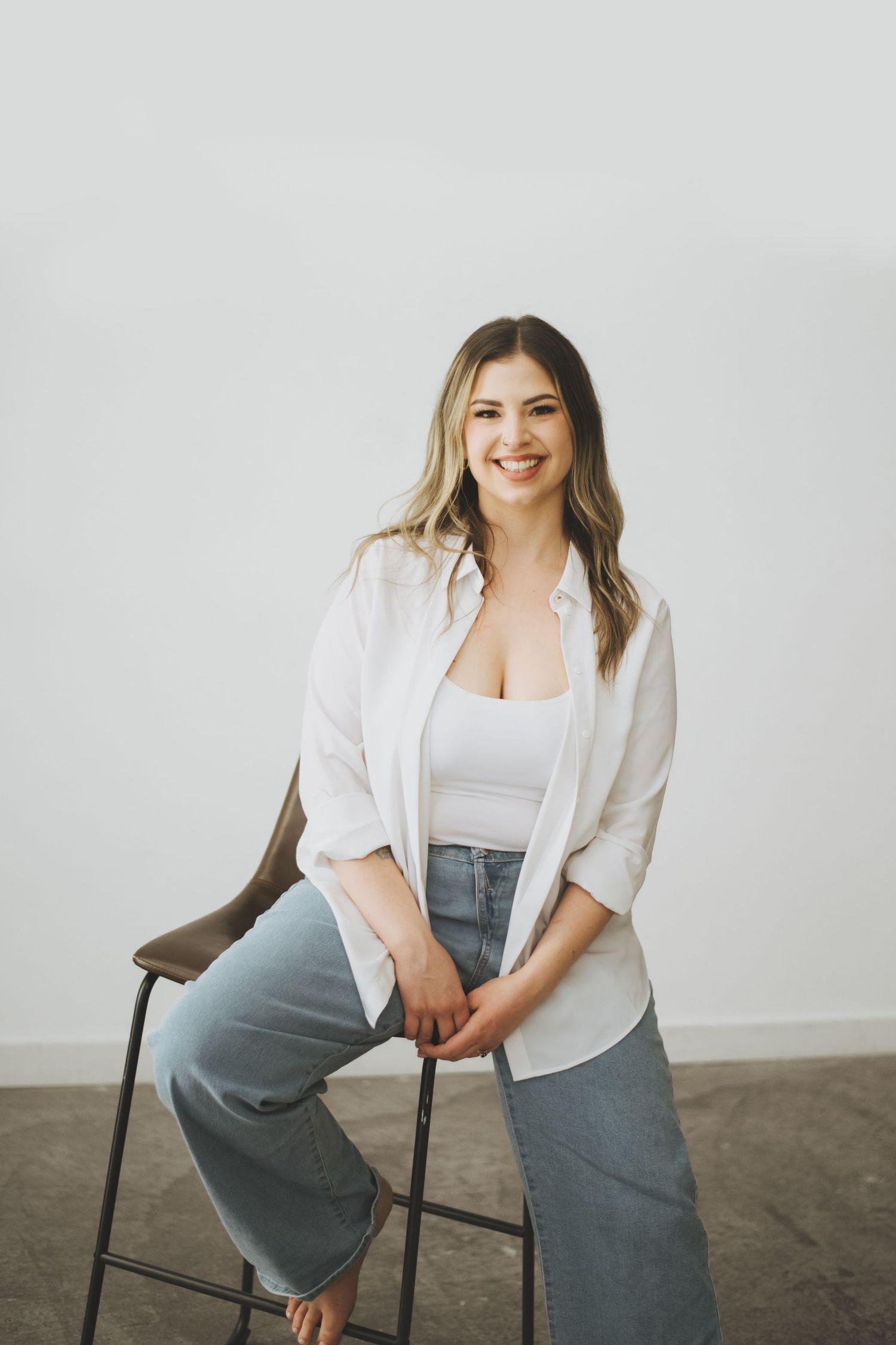 A woman in a white button-down shirt and light blue jeans sits casually on a dark stool against a white background.