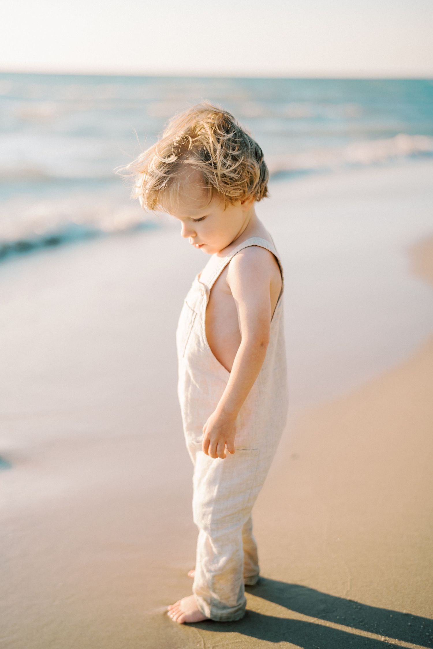 Young child in white clothing standing on a serene beach at sunset while looking down.