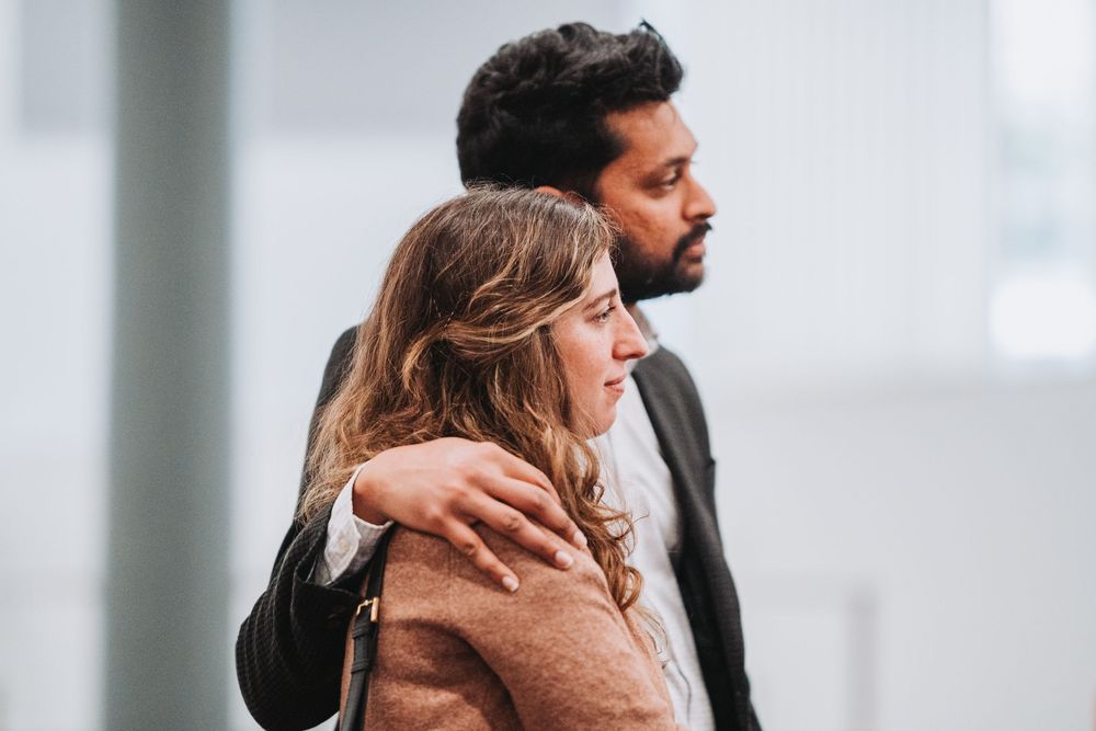 A couple in winter attire stand close together in an intimate embrace against a bright minimalist background.