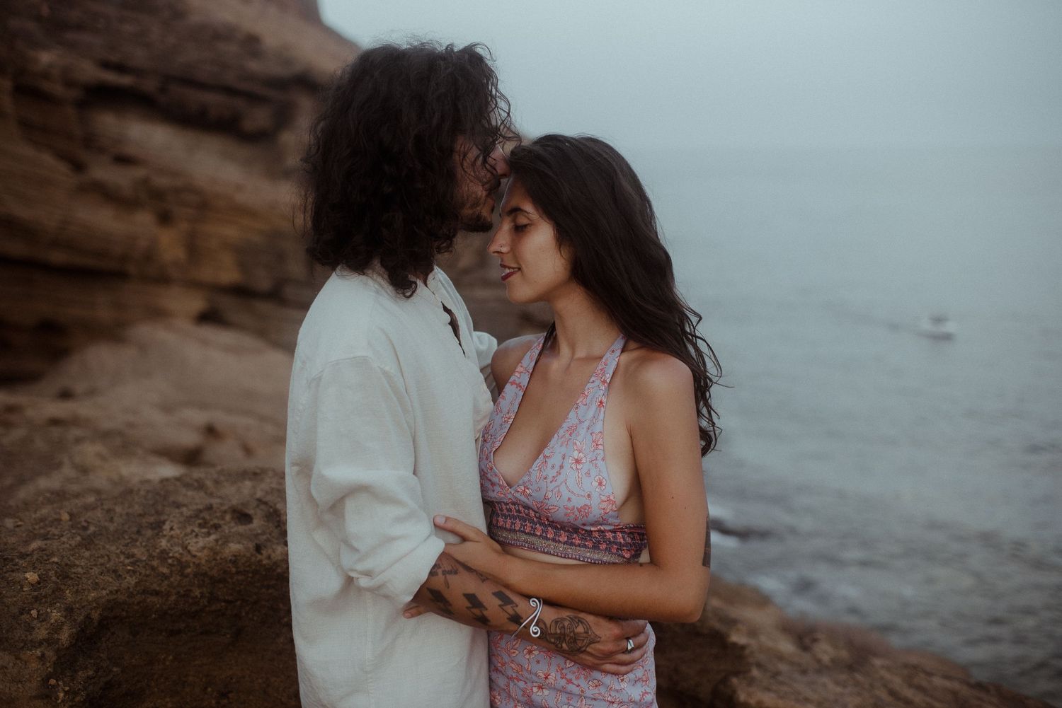 A romantic couple embraces on rocky coastal cliffs during a moody overcast evening.